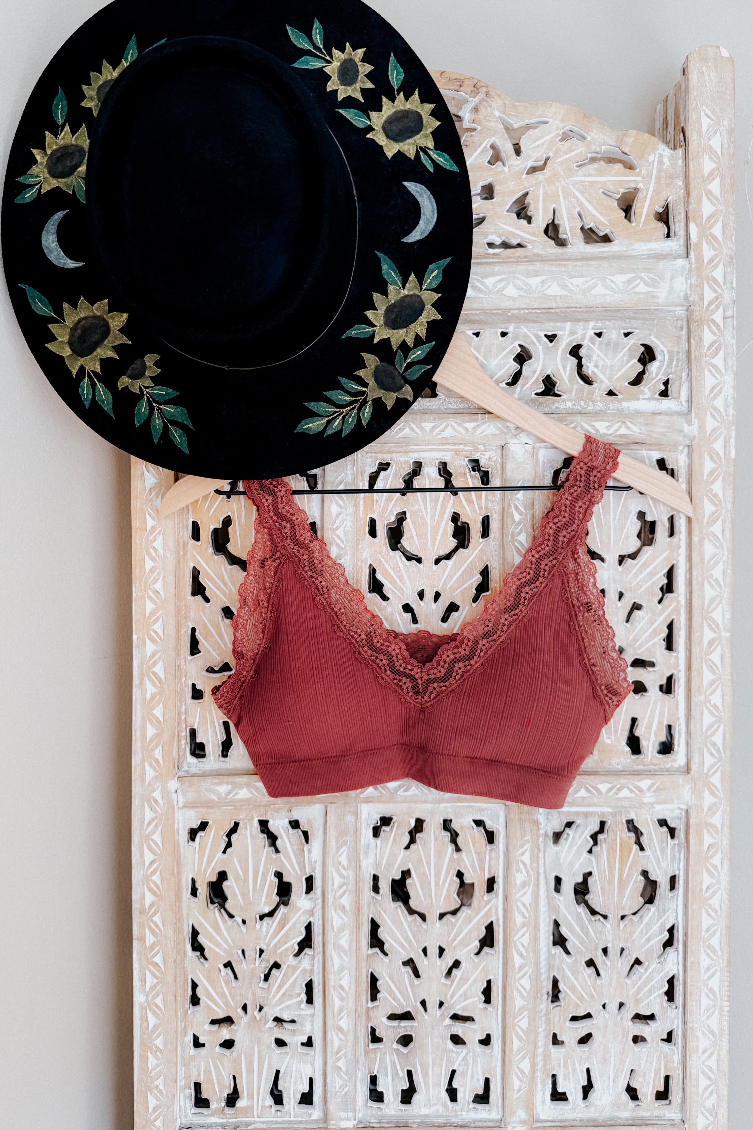 Rust-colored lace-trimmed bralette hanging on decorative carved wooden panel next to black hat with floral embroidery