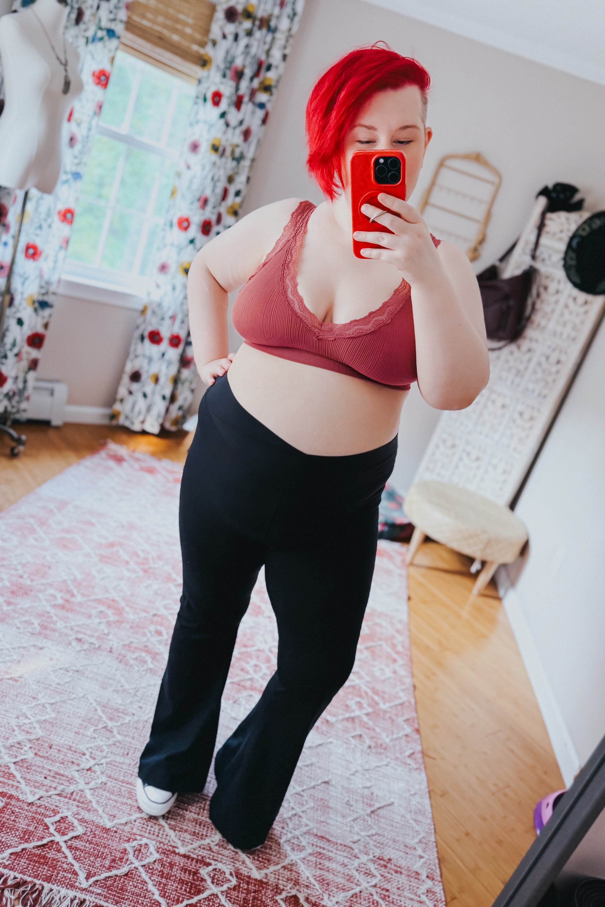 Plus-size woman with bright red hair wearing rust-colored bralette with lace trim and black pants taking mirror selfie in bright room