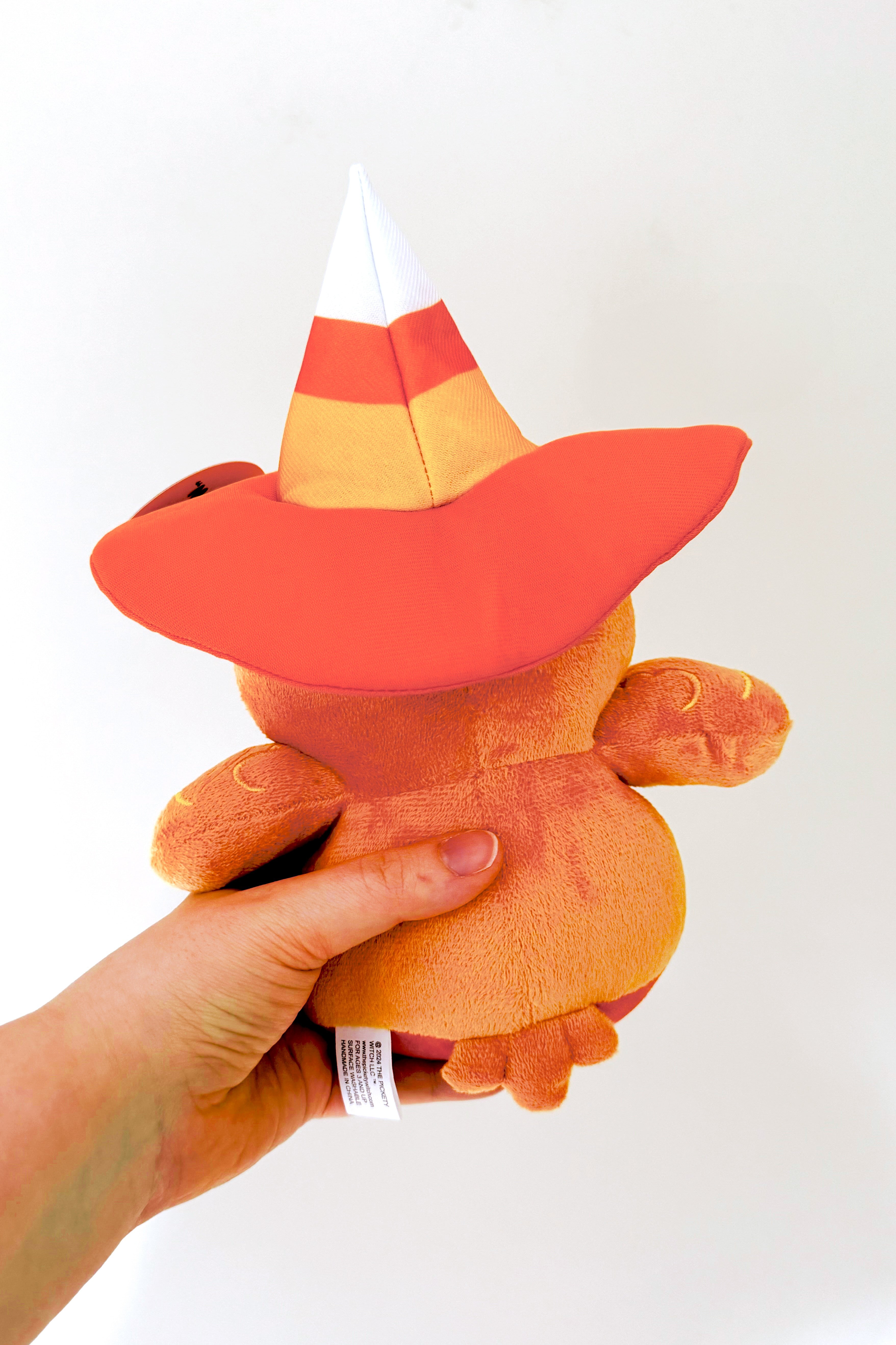 Hand holding a plush pumpkin spice bird seen from behind wearing a candy corn inspired witch hat with wide brim