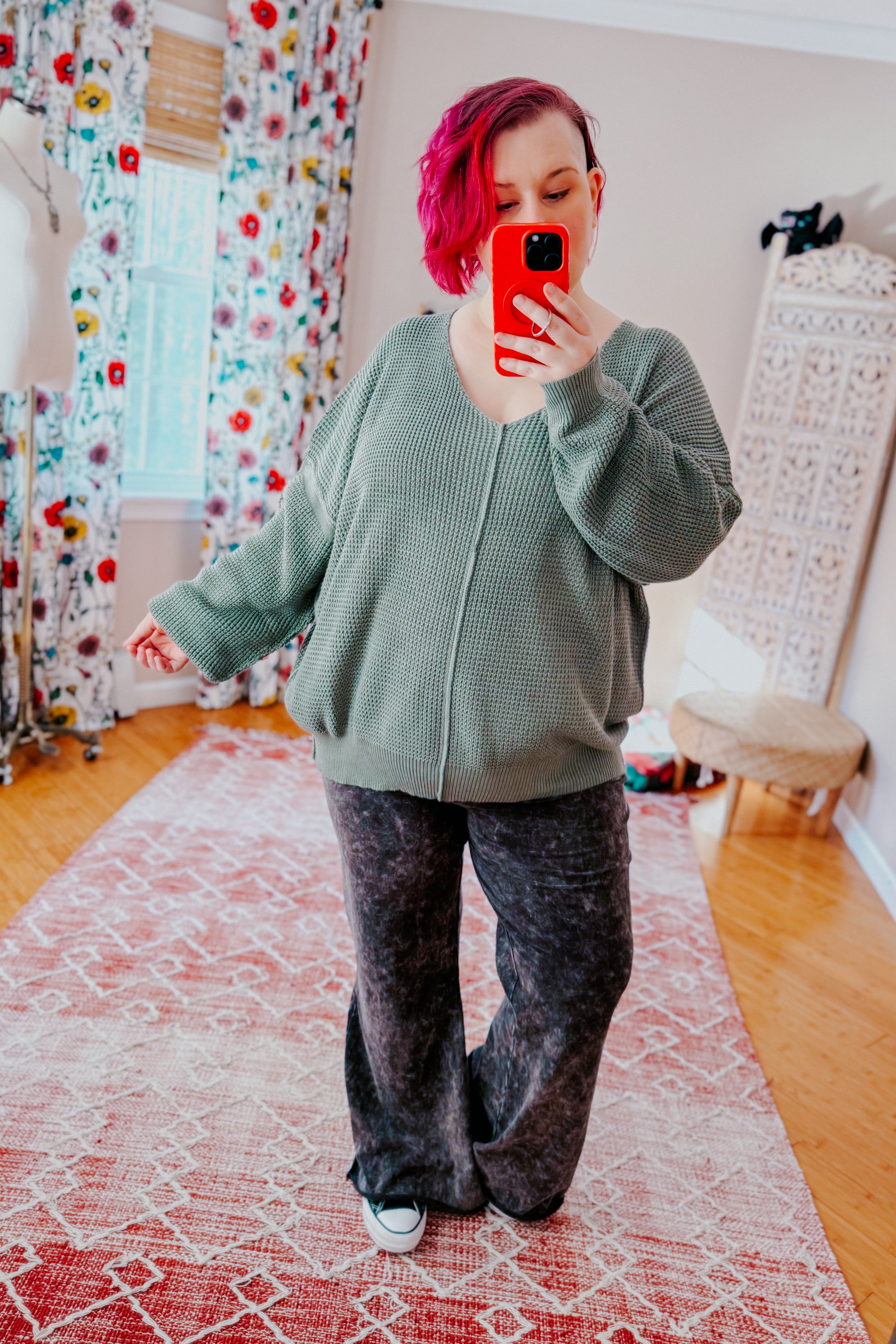 A women with red hair taking a mirror selfie wearing an olive green v-neck sweater with exposed seams, grey acid wash pants, and converse sneakers standing in an indoor setting.