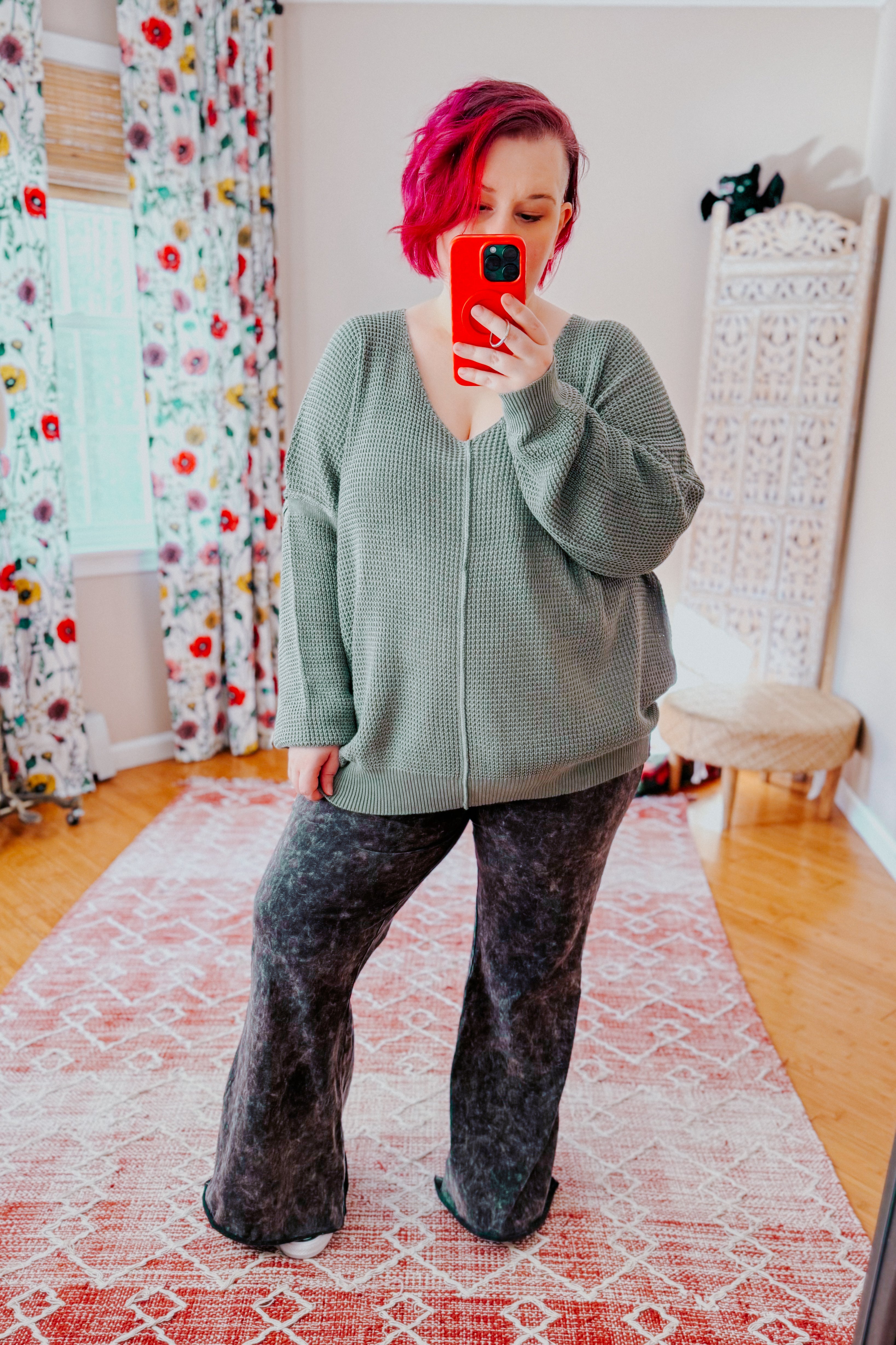 A women with red hair taking a mirror selfie wearing an olive green v-neck sweater with exposed seams, grey acid wash pants, and converse sneakers standing in an indoor setting.