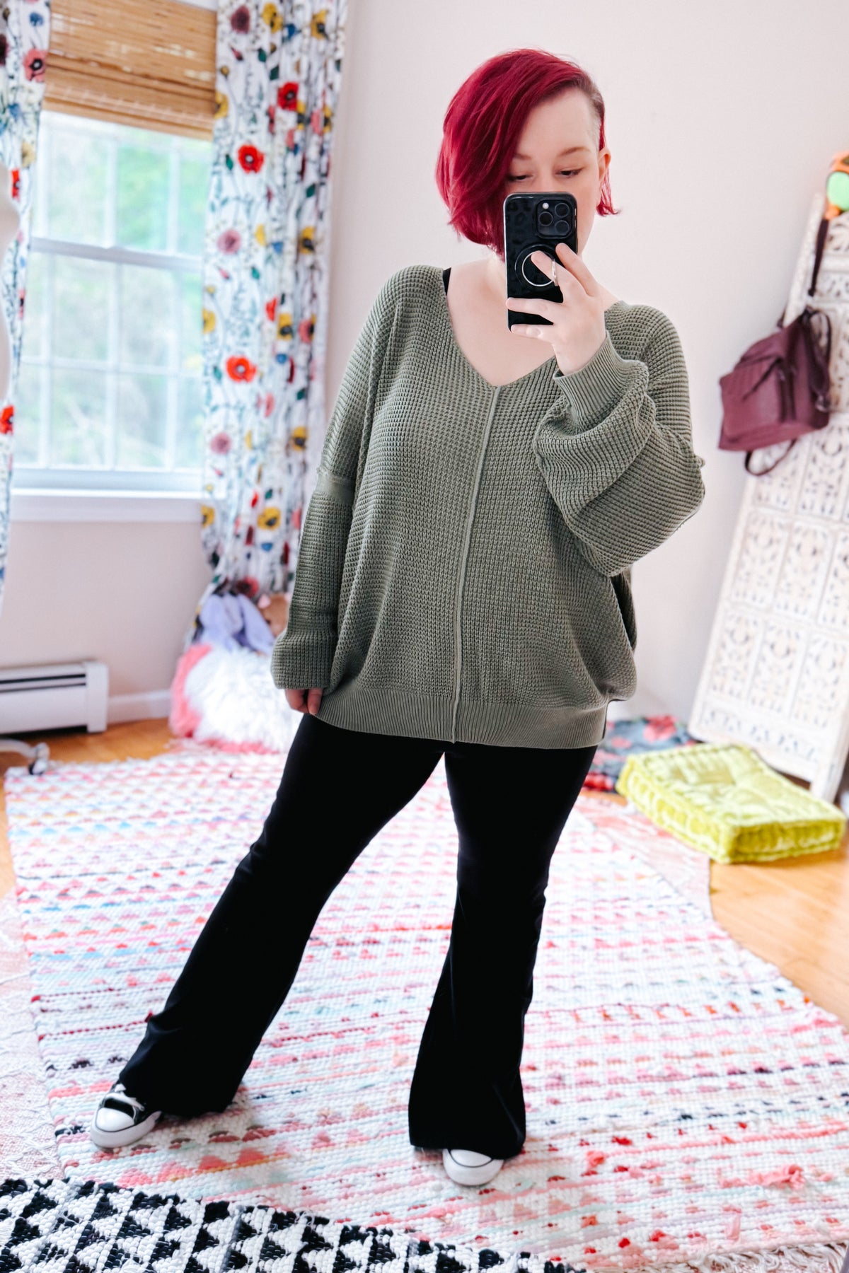 Red haired woman photographing herself in a sage textured V neck sweater, black pants, and white sneakers on a colorful bedroom rug