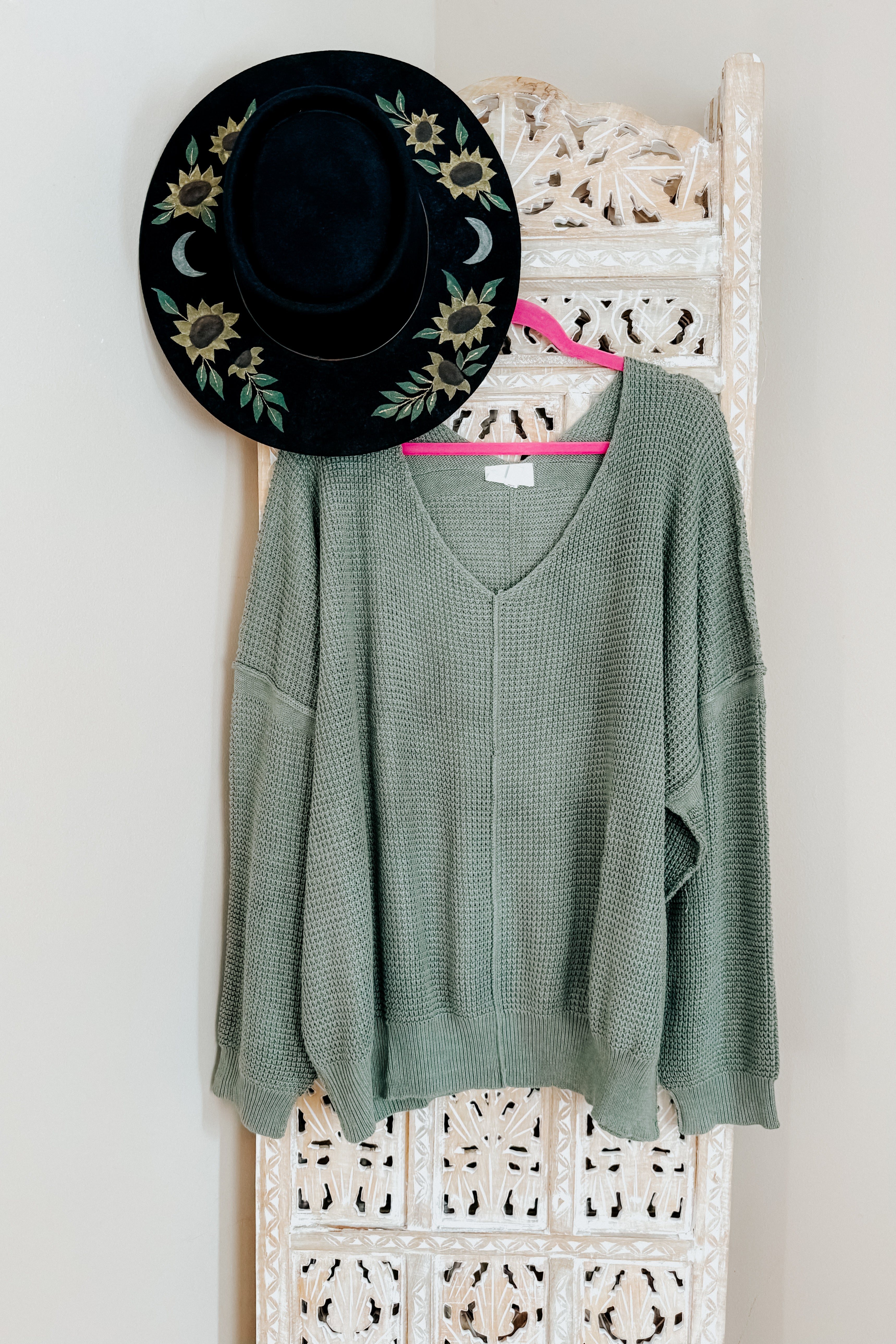 An olive green v-neck sweater with exposed seams hanging from a pink hanger on a ornate room divider with a hand painted wide brim hat.