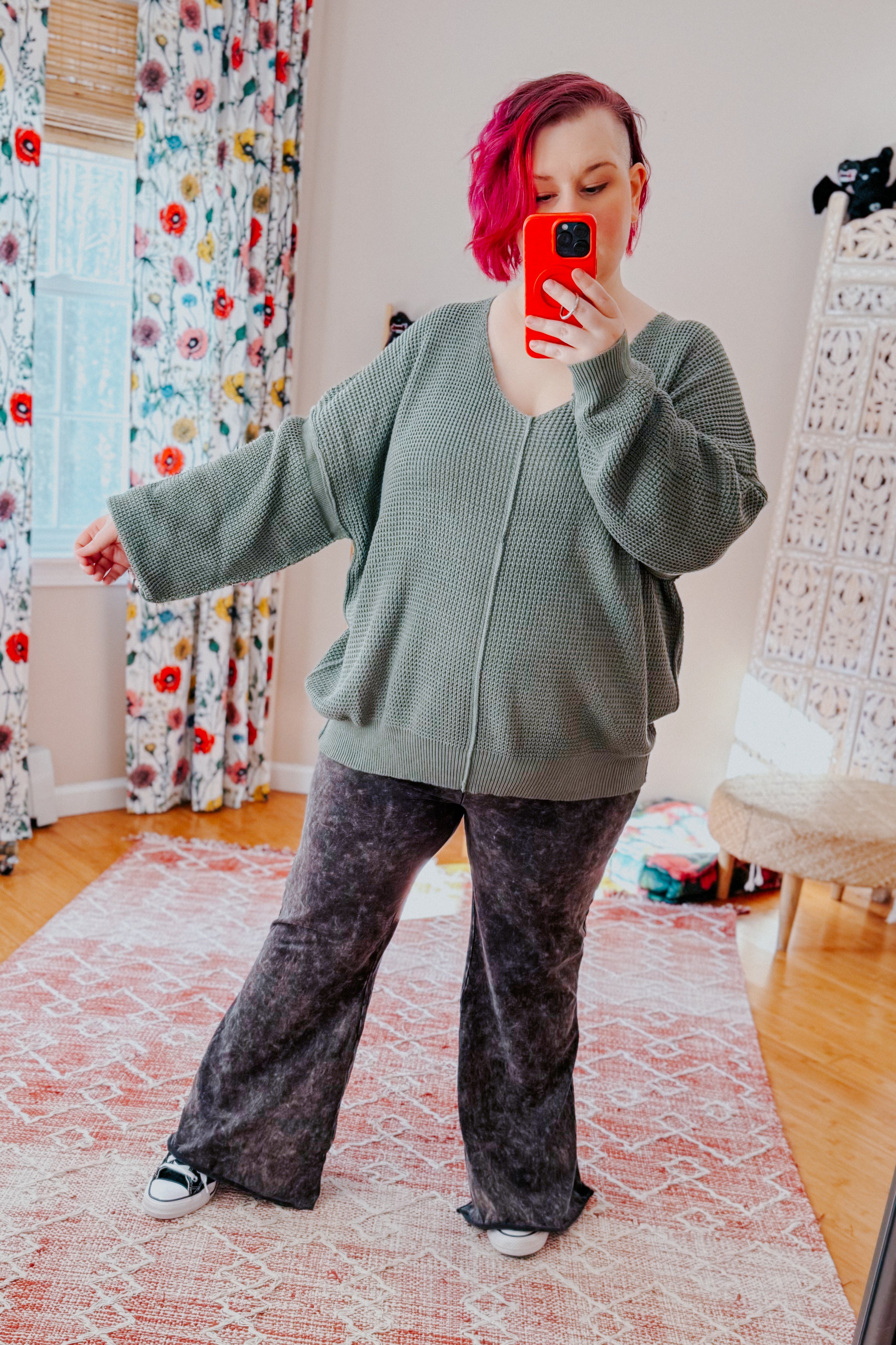 A women with red hair taking a mirror selfie wearing an olive green v-neck sweater with exposed seams, grey acid wash pants, and converse sneakers standing in an indoor setting.