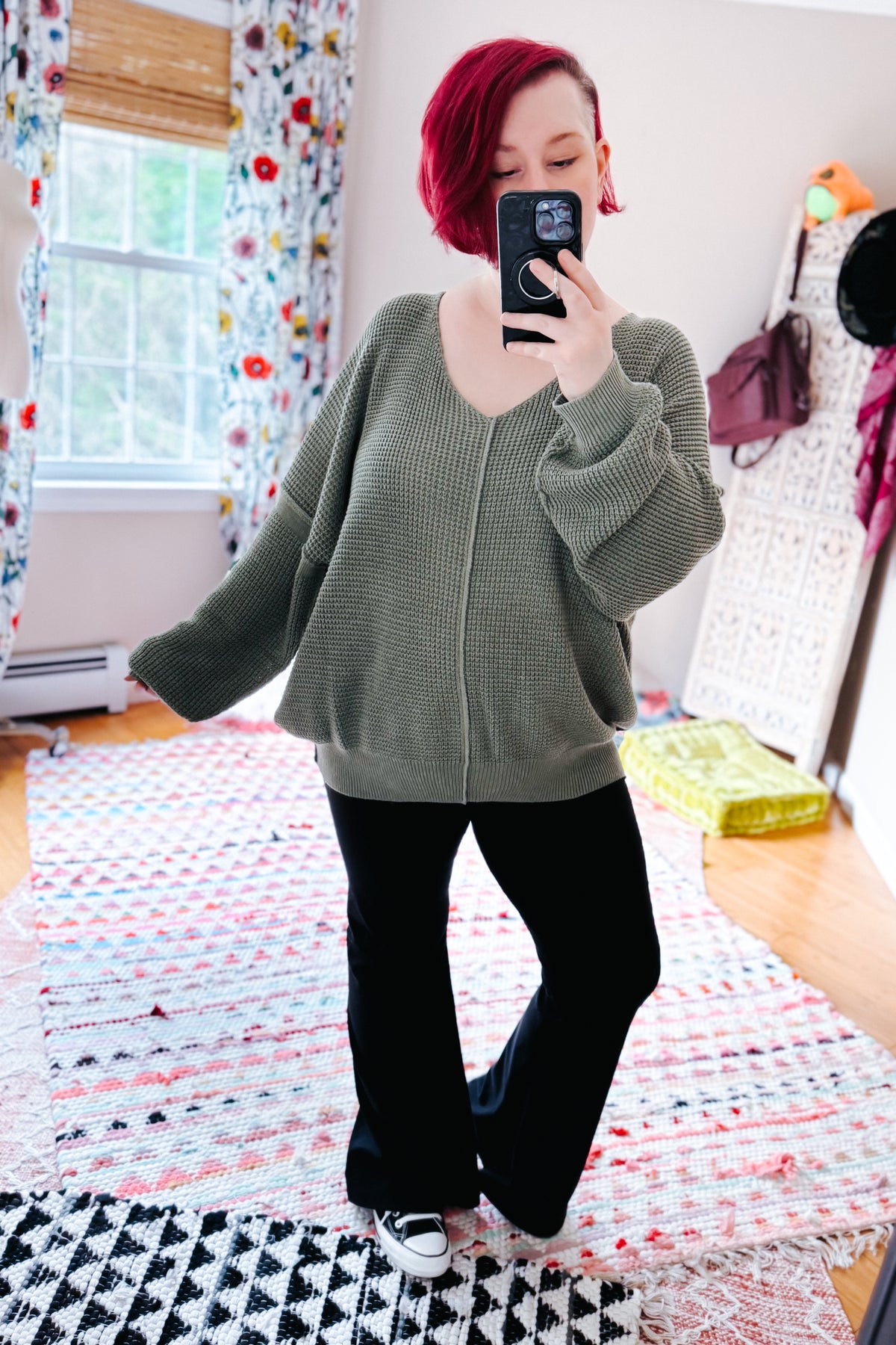 Red haired woman posing for a bedroom mirror selfie in a sage V neck sweater with black pants and sneakers on layered rugs