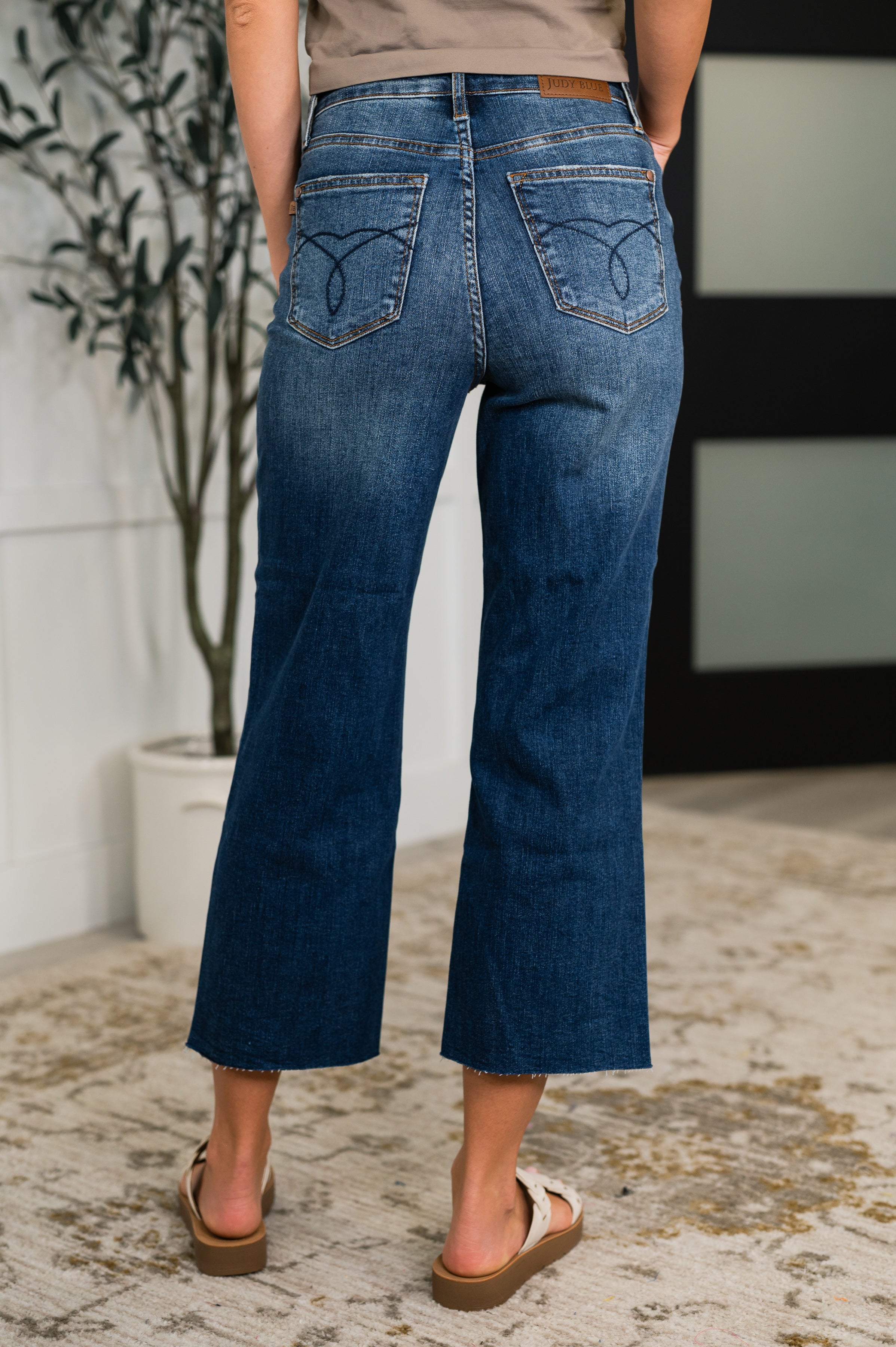 Blue mid-rise denim cropped flare jeans with stitched back pockets and raw hem worn with beige sandals indoors
