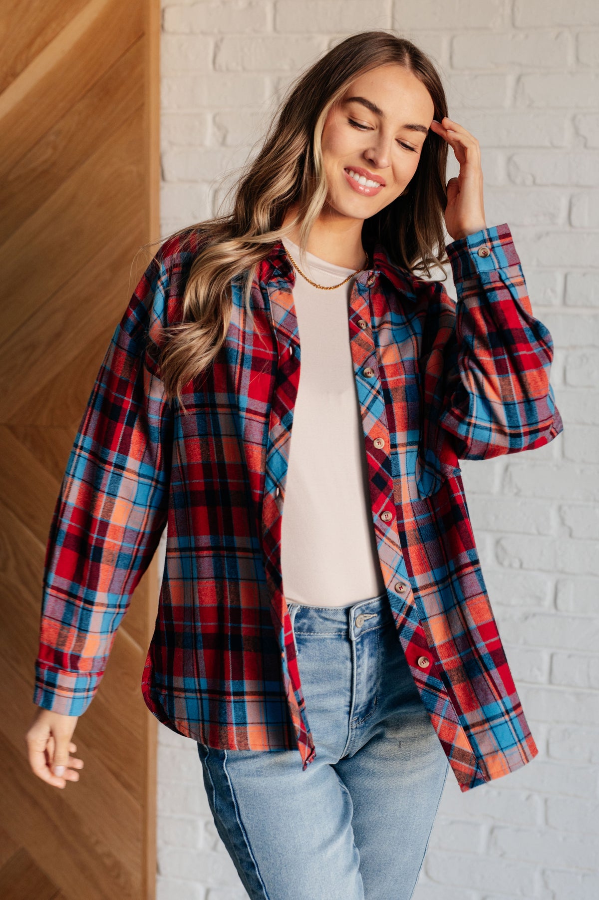 Woman wearing a red plaid button up shirt over a white top with jeans while posing beside a white brick wall