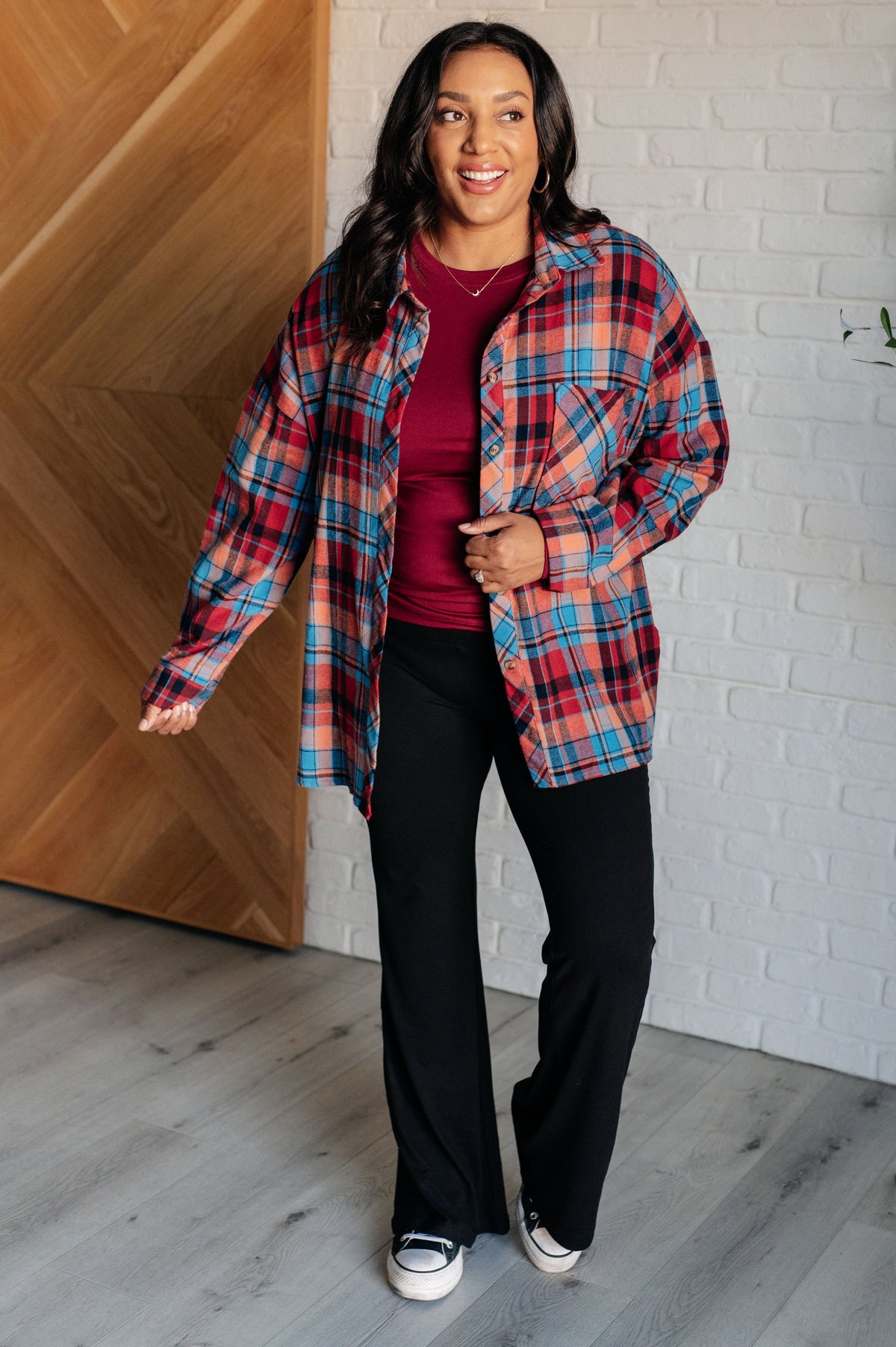 Smiling woman wearing a red plaid button up over a burgundy top with black pants and sneakers indoors beside a white brick wall
