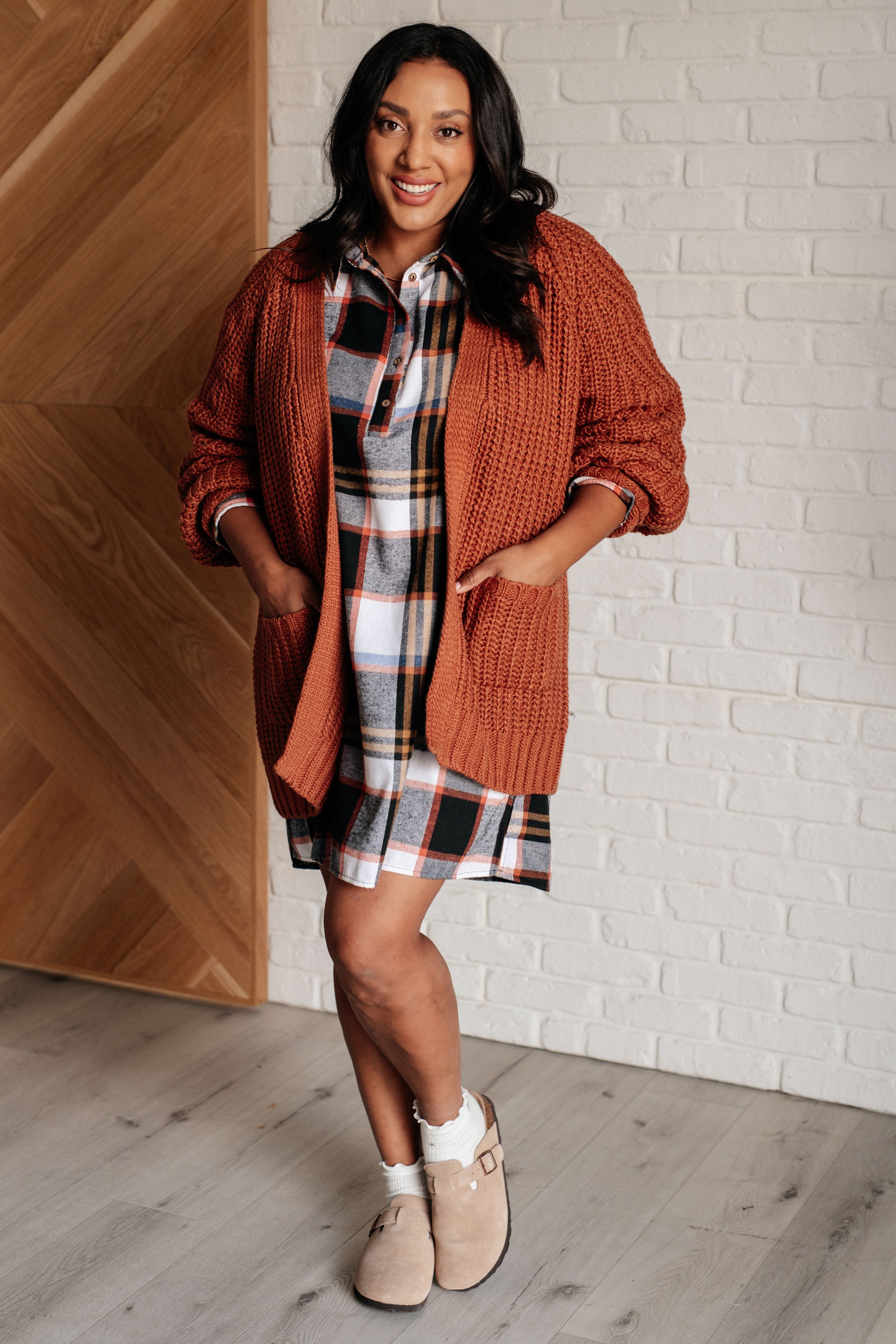 Woman wearing a plaid dress layered with a rust knit cardigan and beige slip-on shoes indoors