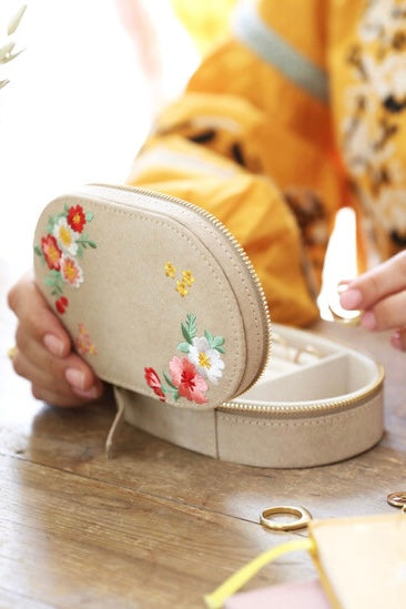 Hand holding cream velvet jewelry box with floral embroidery and gold zipper on wooden table