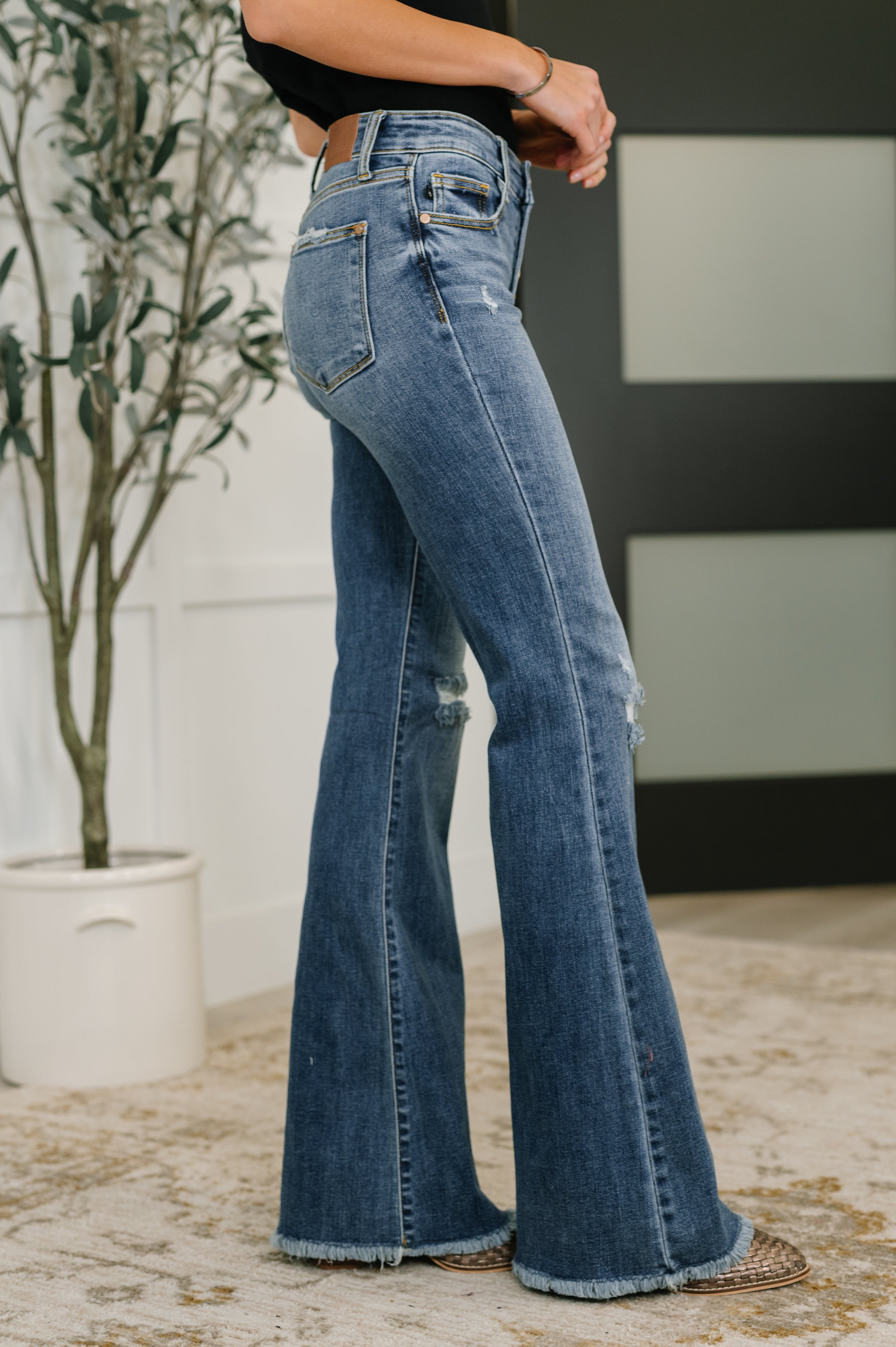 Side view of mid-rise blue denim flare jeans with distressed frayed hems worn indoors
