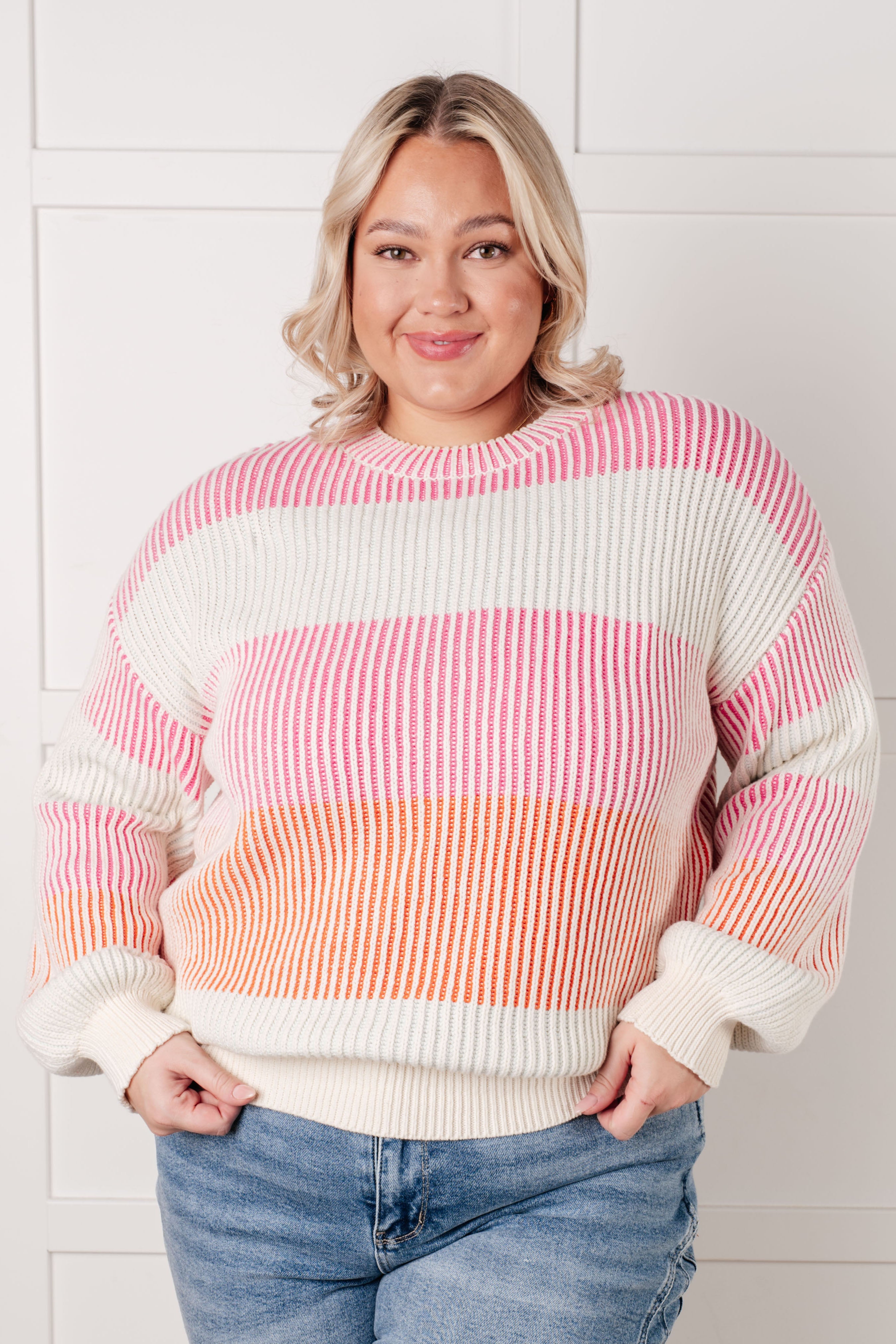 Smiling woman indoors wearing a ribbed striped top in pink, white, and orange paired with light wash jeans