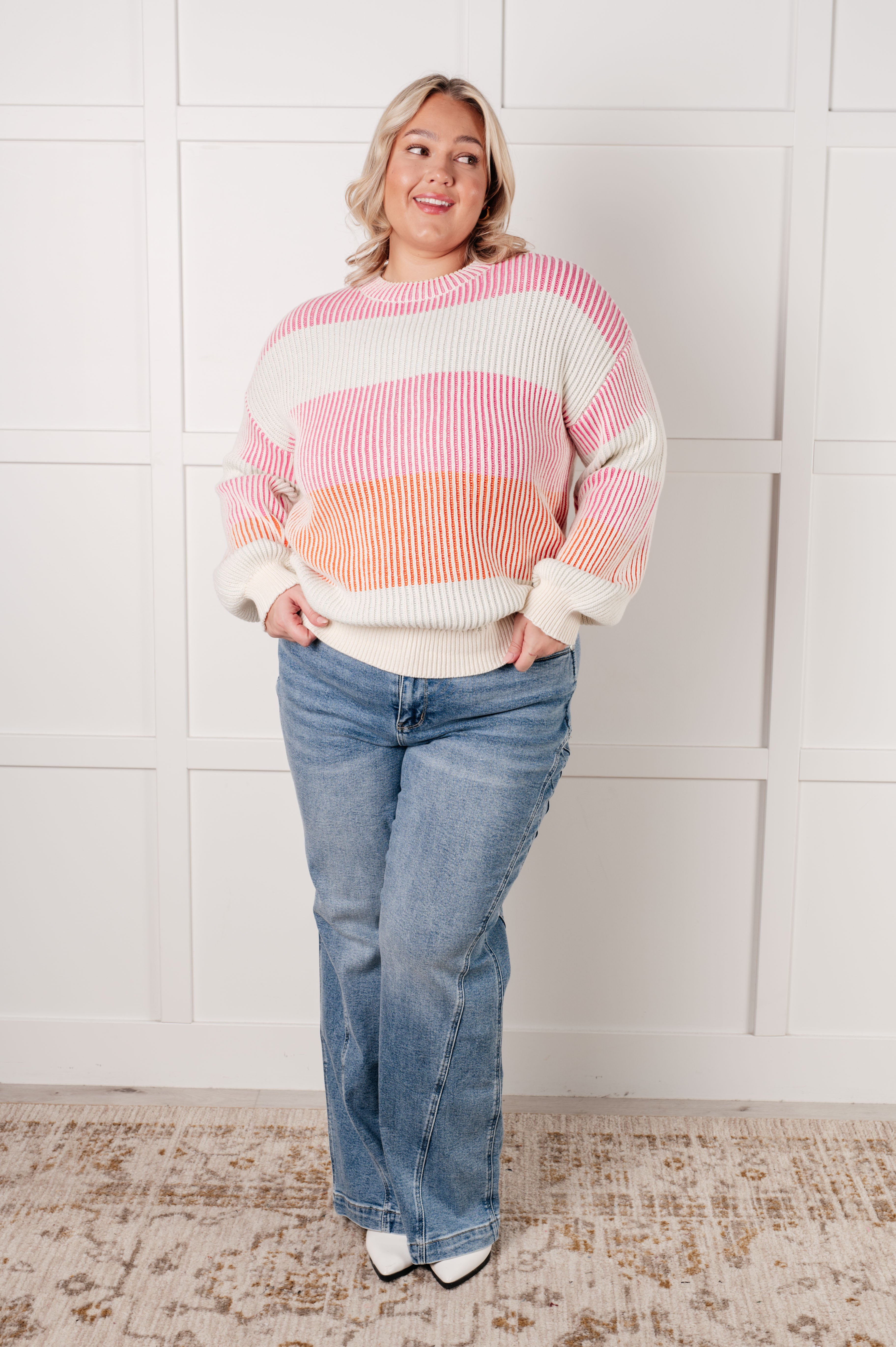 Woman wearing a pink and orange striped ribbed sweater paired with blue straight-leg jeans and white shoes indoors