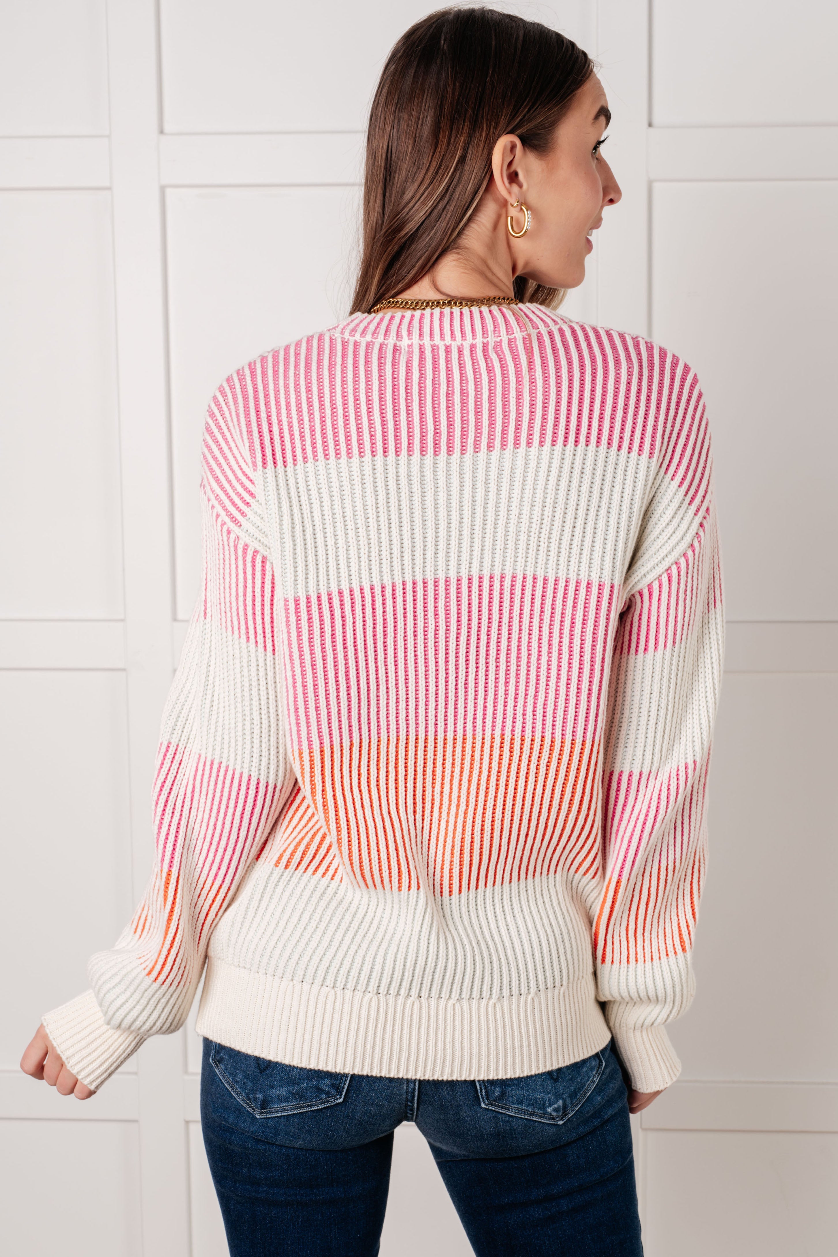 Woman seen from behind wearing a ribbed striped top in pink, orange, and cream tones paired with dark blue jeans indoors