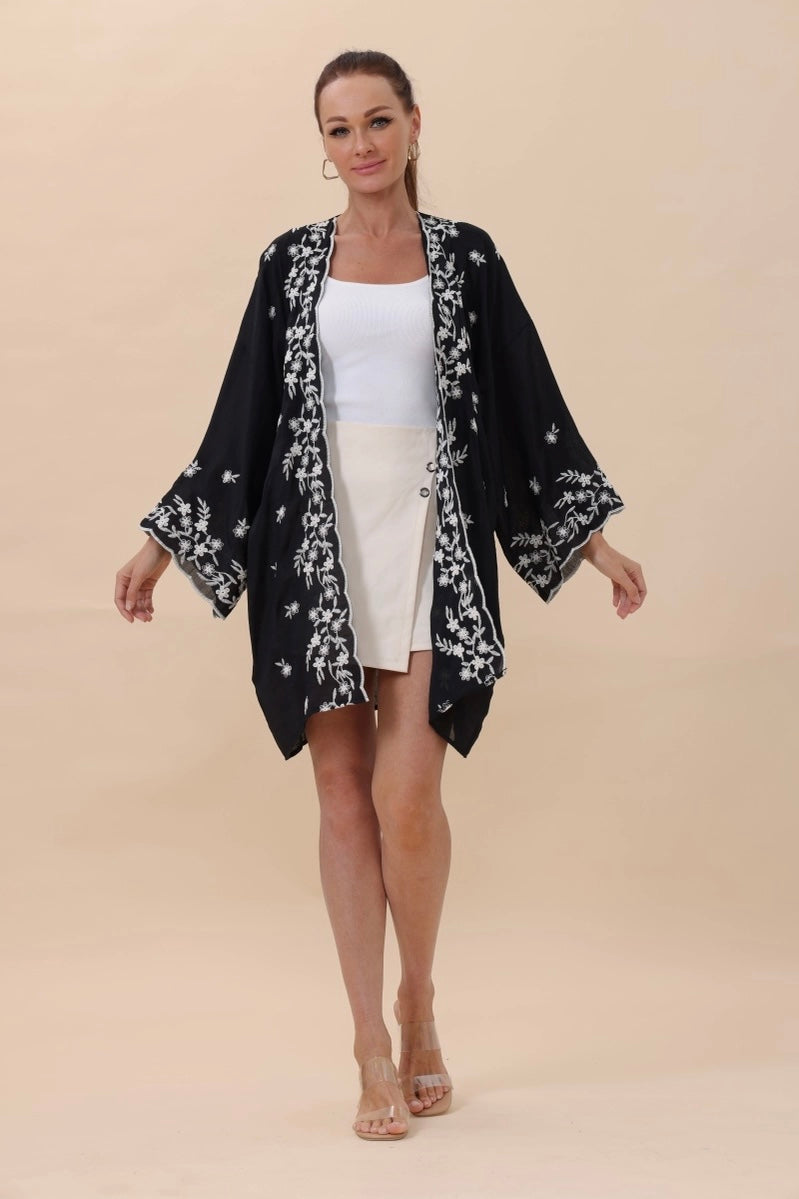 Woman wearing a black duster with white floral embroidery over a white tank top and cream skirt against beige background