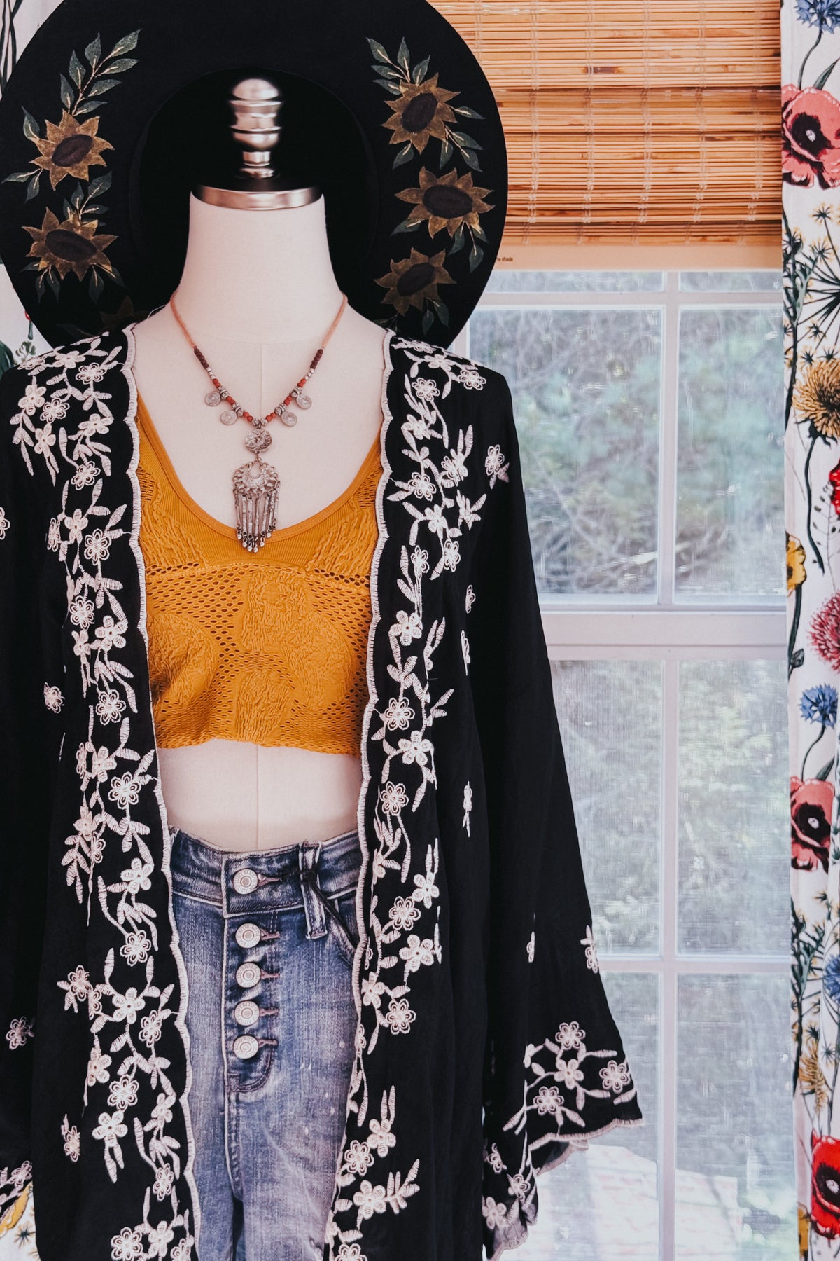 Black floral embroidered duster layered over a mustard knit crop top and high waisted jeans on a mannequin indoors