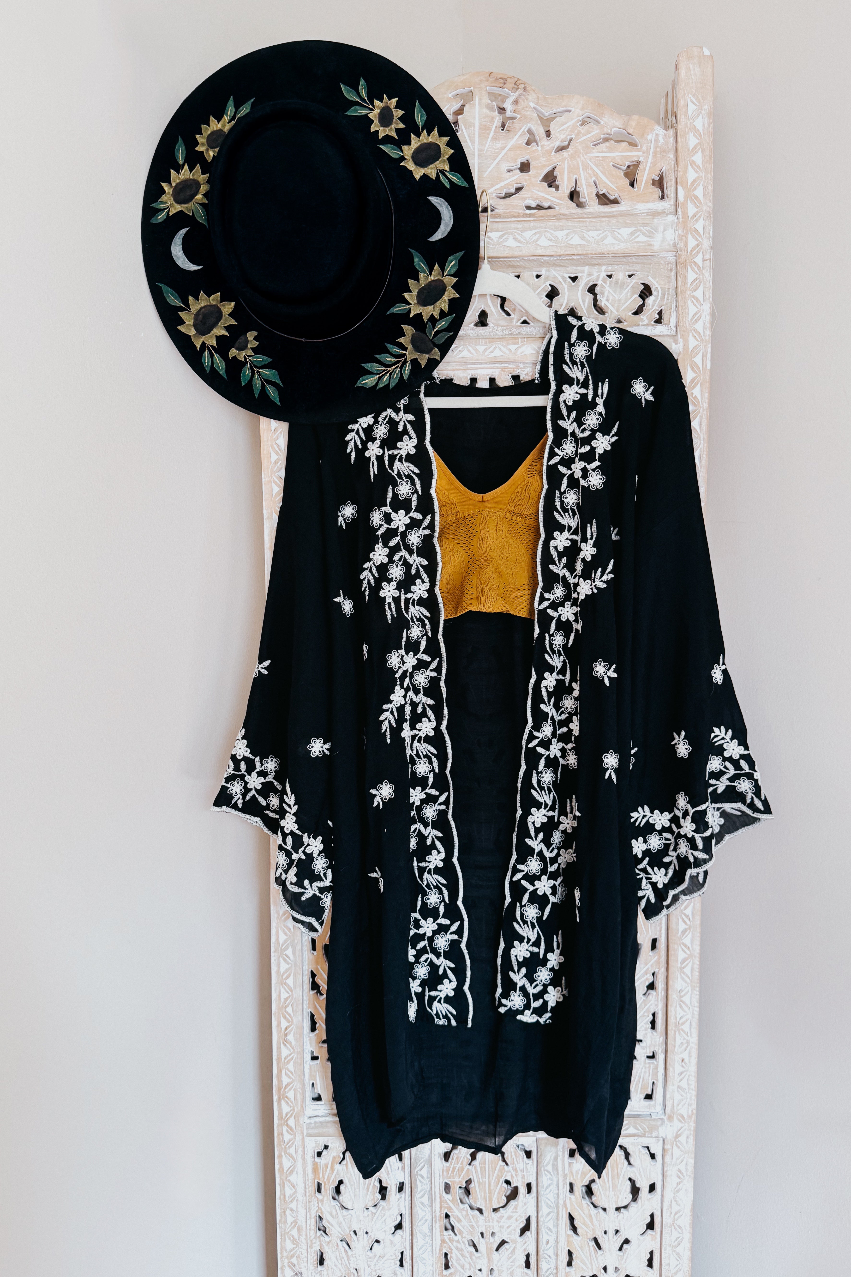 Black duster with white floral embroidery hanging on carved wooden panel with black embroidered hat and mustard top inside