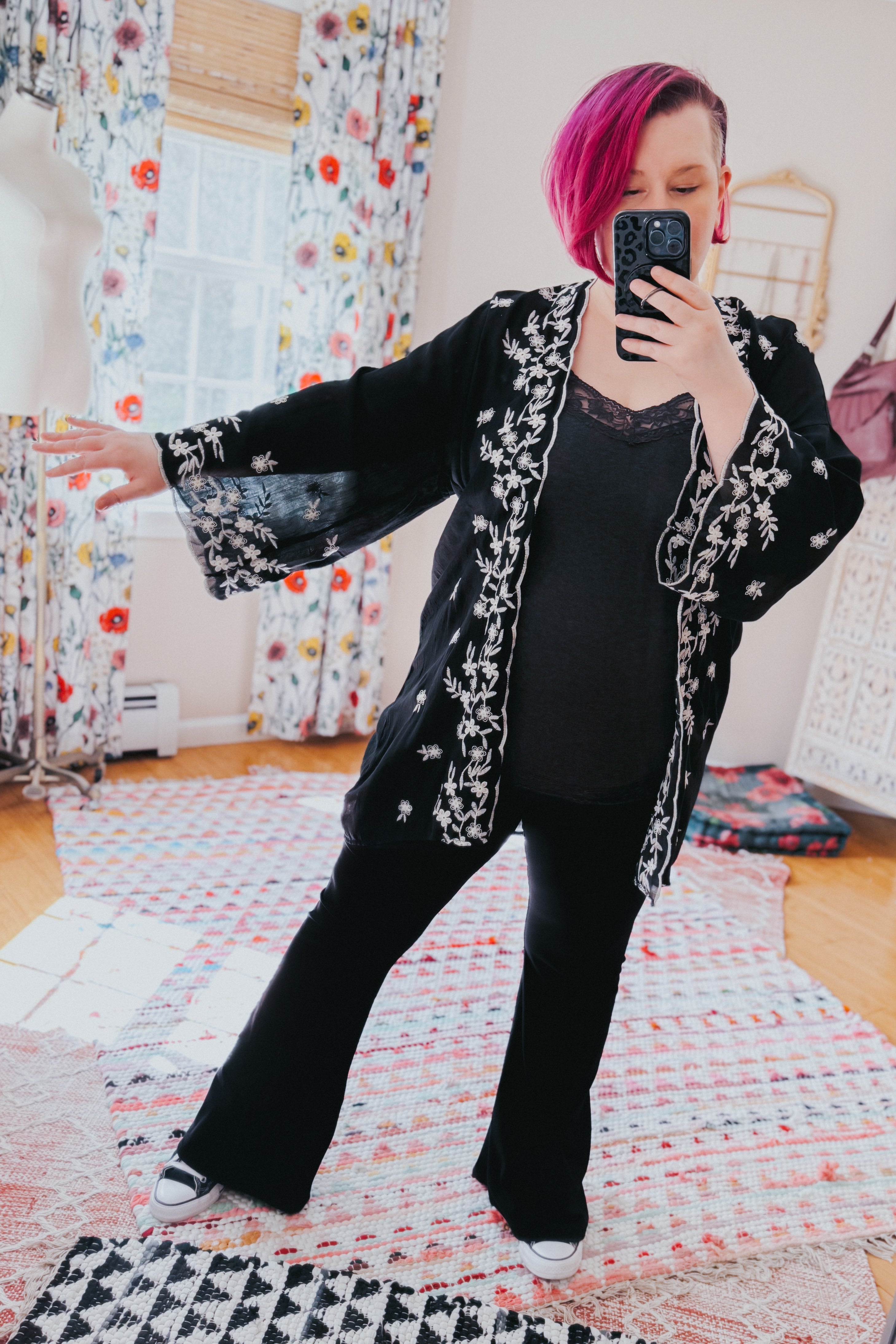 Woman taking mirror selfie wearing a black duster with white floral embroidery over dark top and flared pants indoors