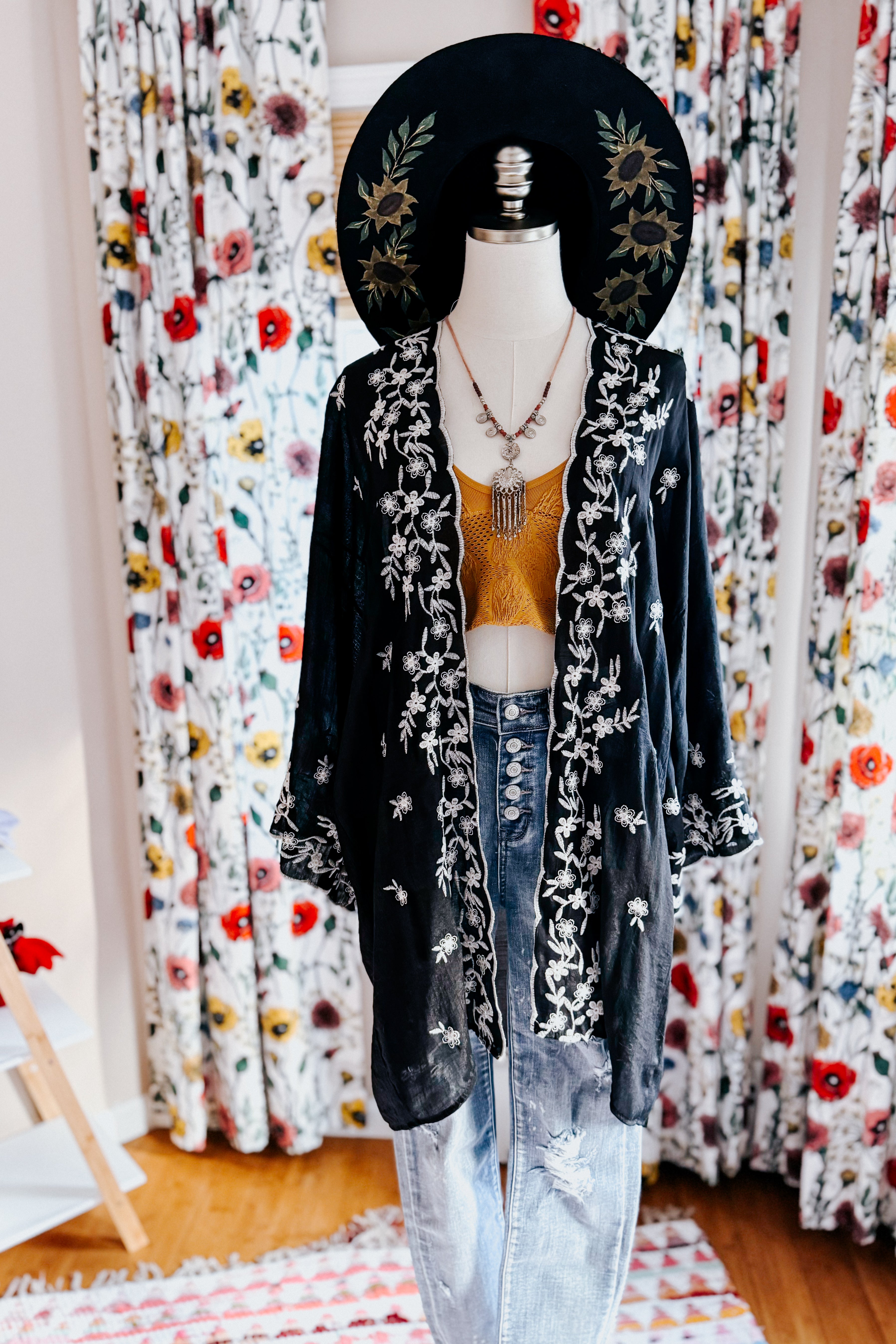 Mannequin dressed in black floral embroidered duster layered over mustard knit top and high-waisted light blue jeans with distressed knees