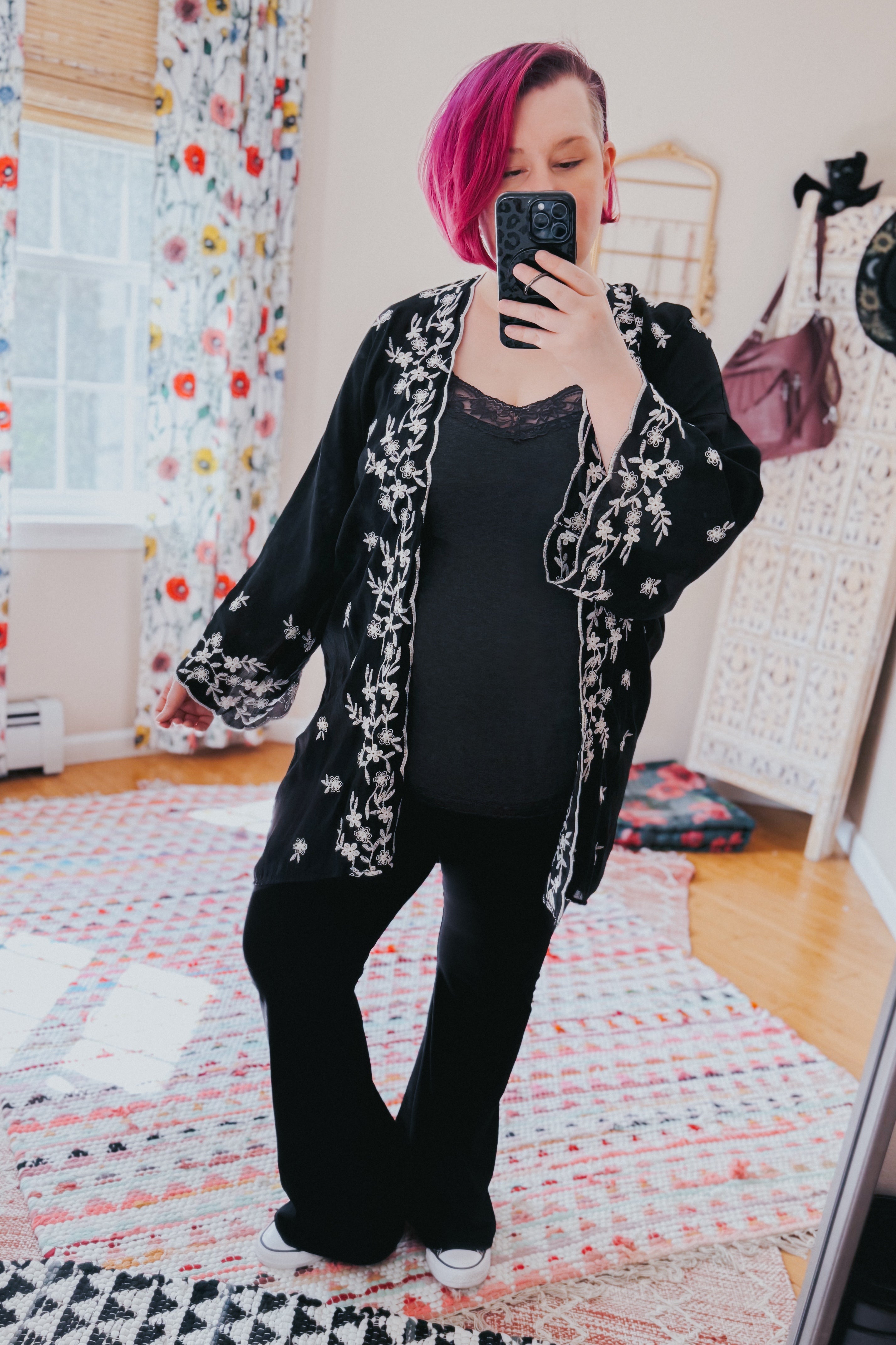 Woman taking selfie wearing black floral embroidered duster over black top and pants in colorful room with patterned rug