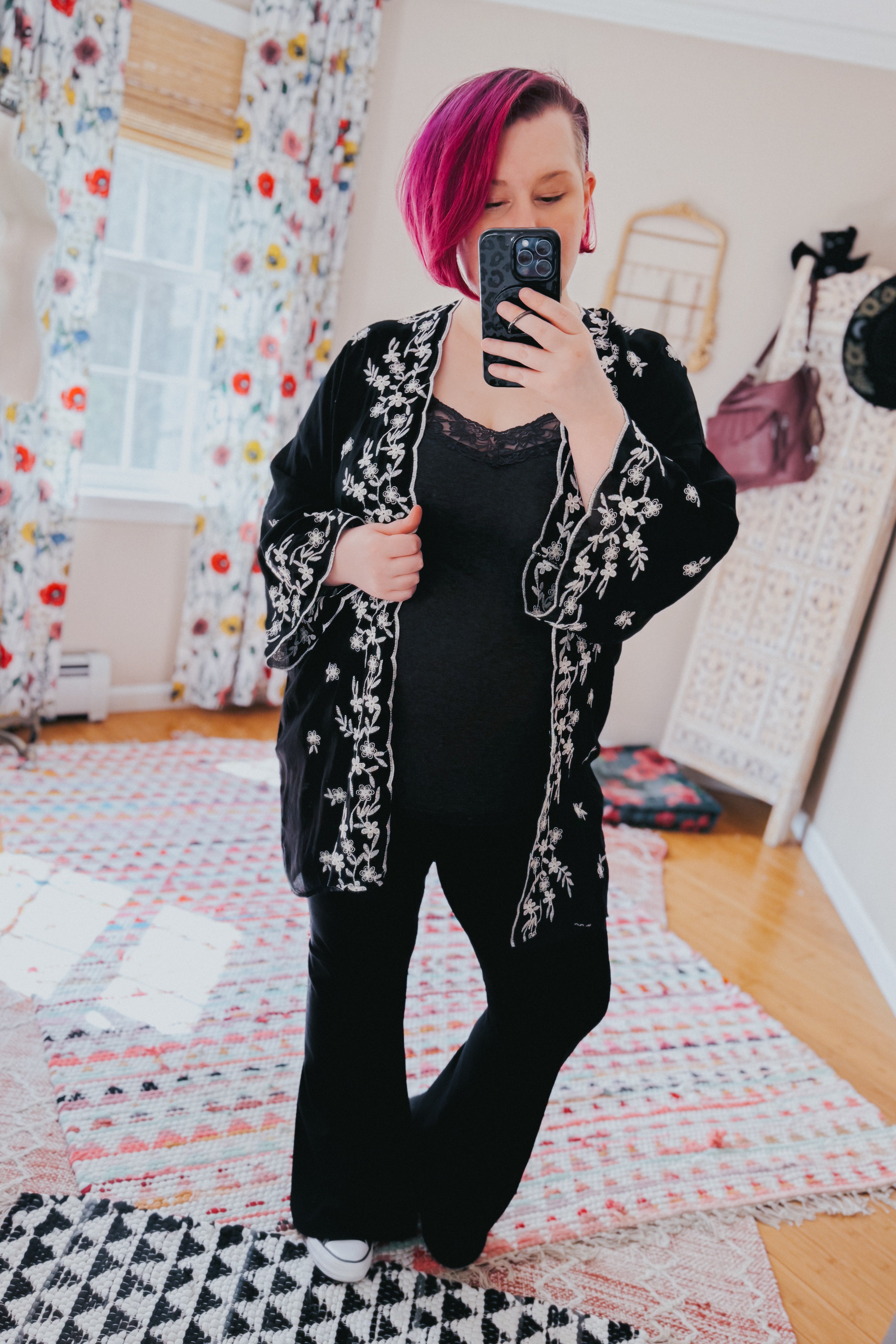 Woman wearing a black floral embroidered duster over black lace-trimmed top and flared pants taking a mirror selfie indoors