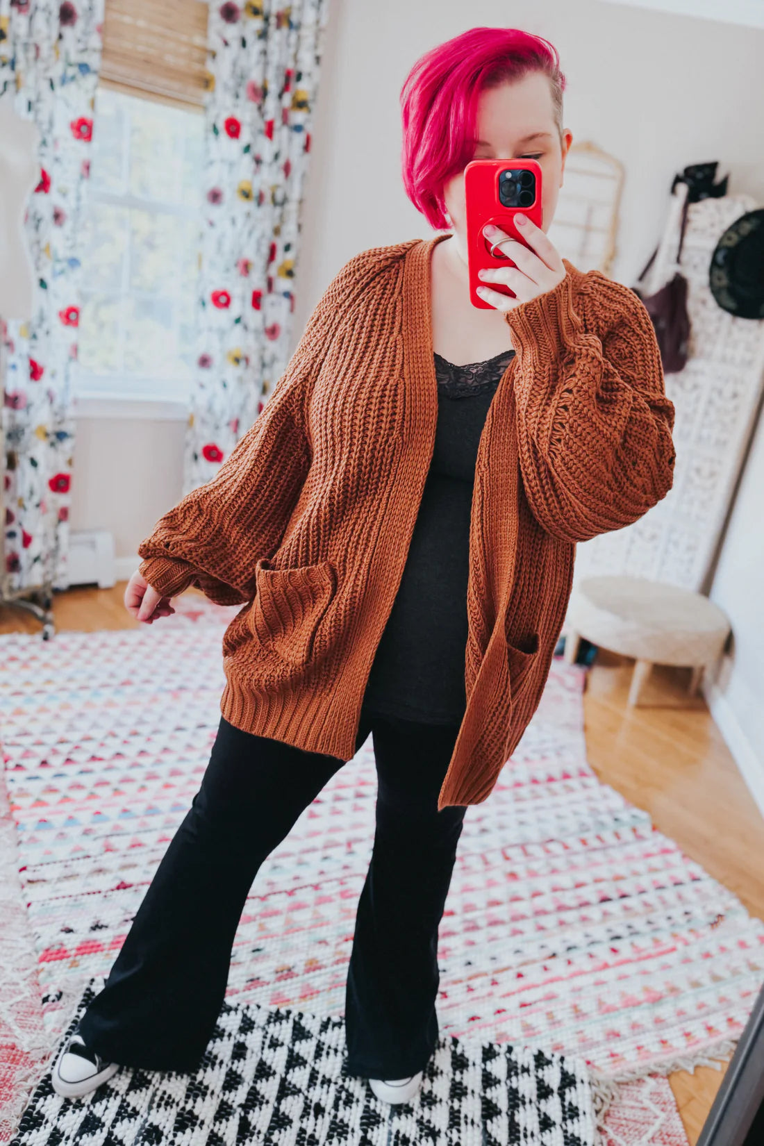 Woman with bright pink hair wearing a chestnut chunky knit cardigan with pockets over a black lace-trimmed top and black pants indoors