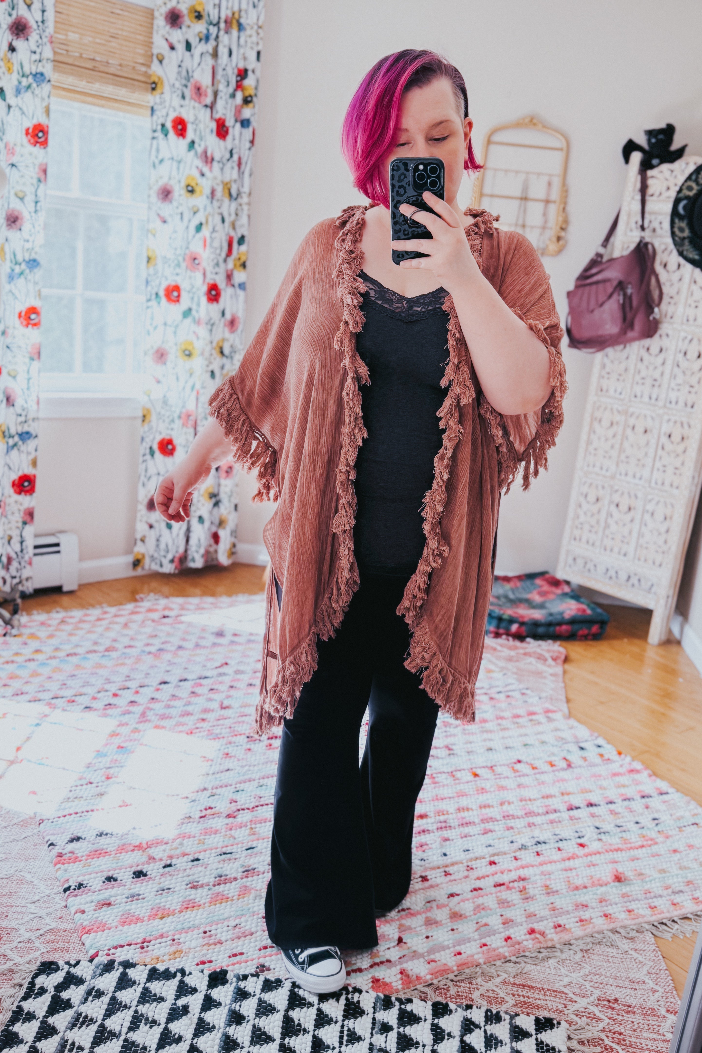 Woman with magenta hair wearing a rose-colored fringe duster over a black outfit taking a mirror selfie indoors