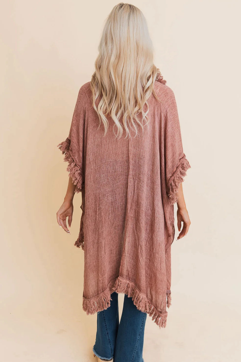 Woman with long blonde hair wearing a rose-colored fringed duster over blue jeans against a neutral background