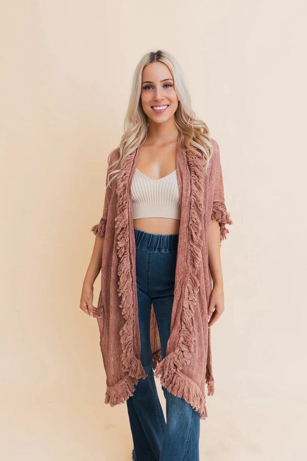 Woman wearing a sunbleached rose fringe duster over cream crop top and high-waisted blue jeans with a relaxed smile
