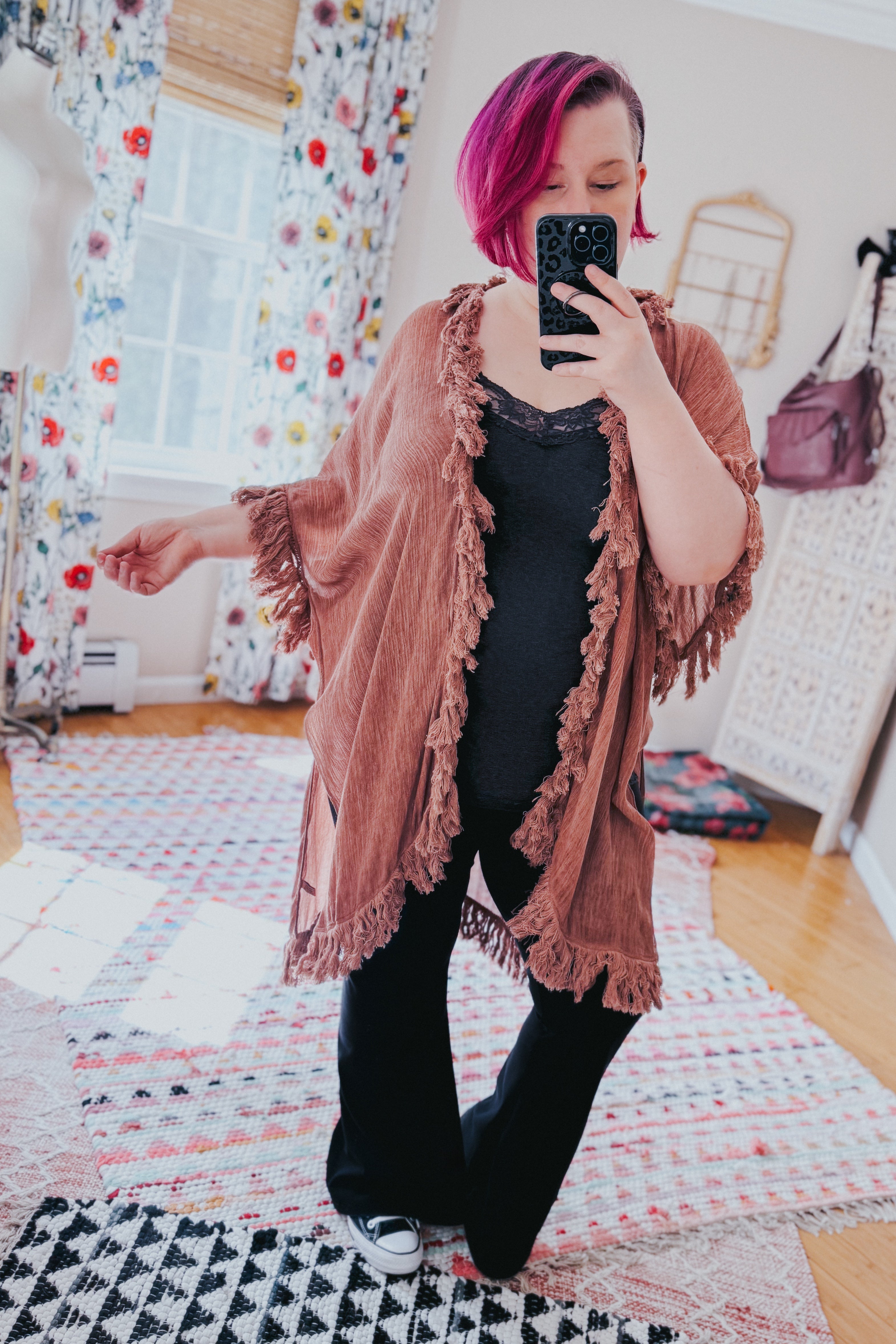 Woman with short magenta hair wearing a rose-colored fringed duster over black outfit in bright eclectic room
