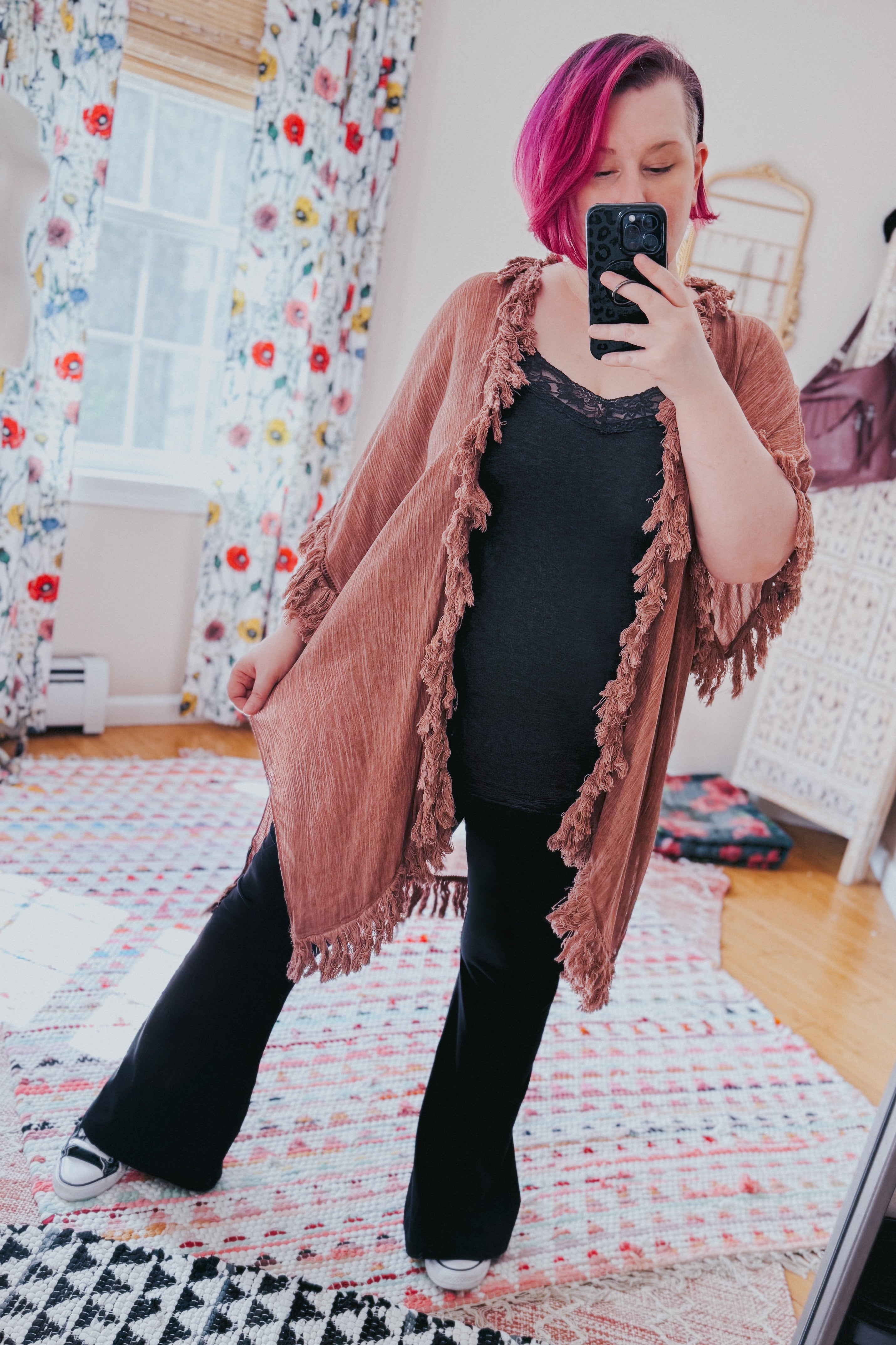 Woman with pink hair wearing a rose-colored fringe duster over black outfit, taking a mirror selfie in a bright room