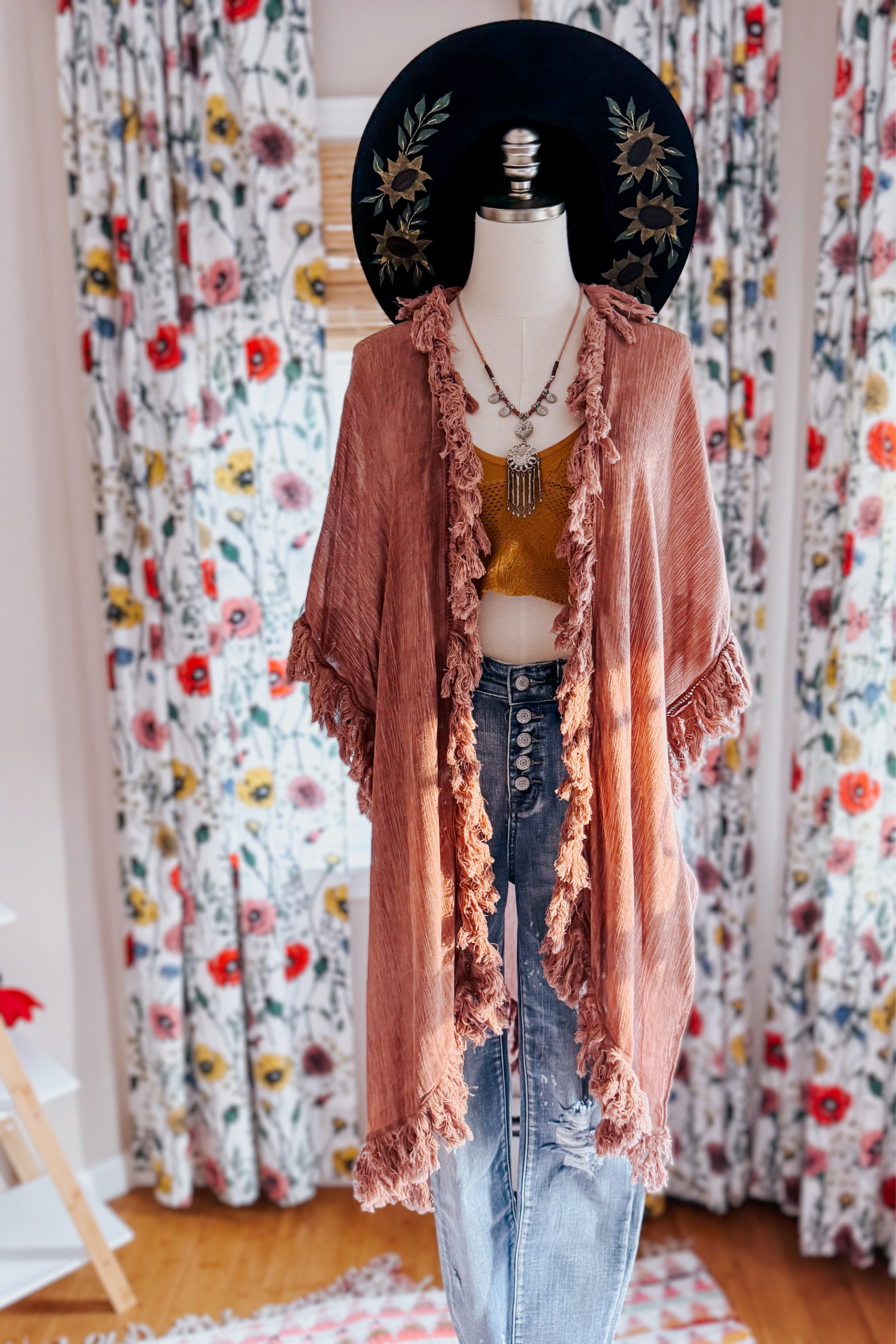 Mannequin wearing rose-colored fringe duster over mustard top and high-waisted button-fly jeans with floral hat indoors