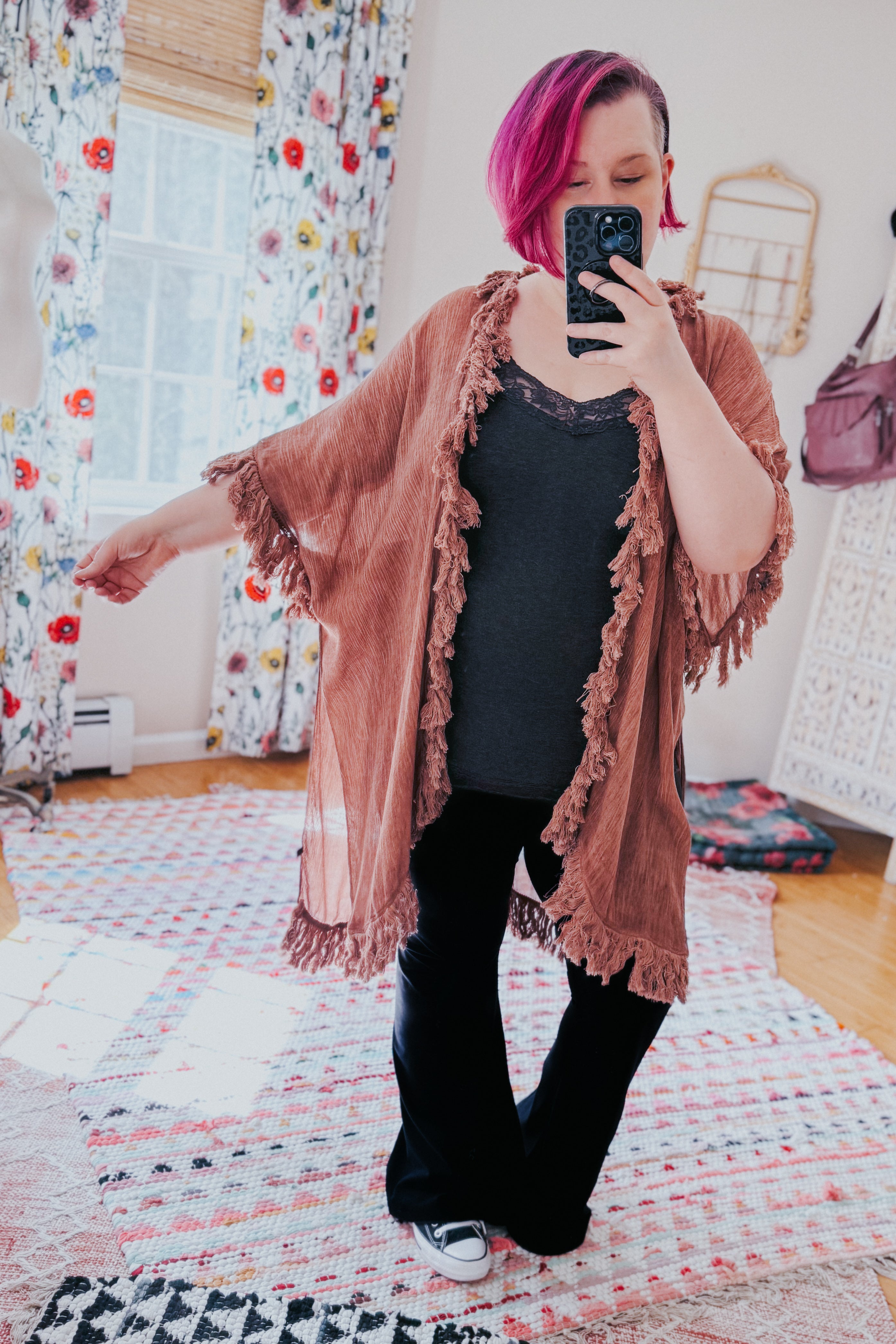Woman with magenta hair wearing a rose-colored fringed duster over dark casual outfit taking a selfie in cozy room