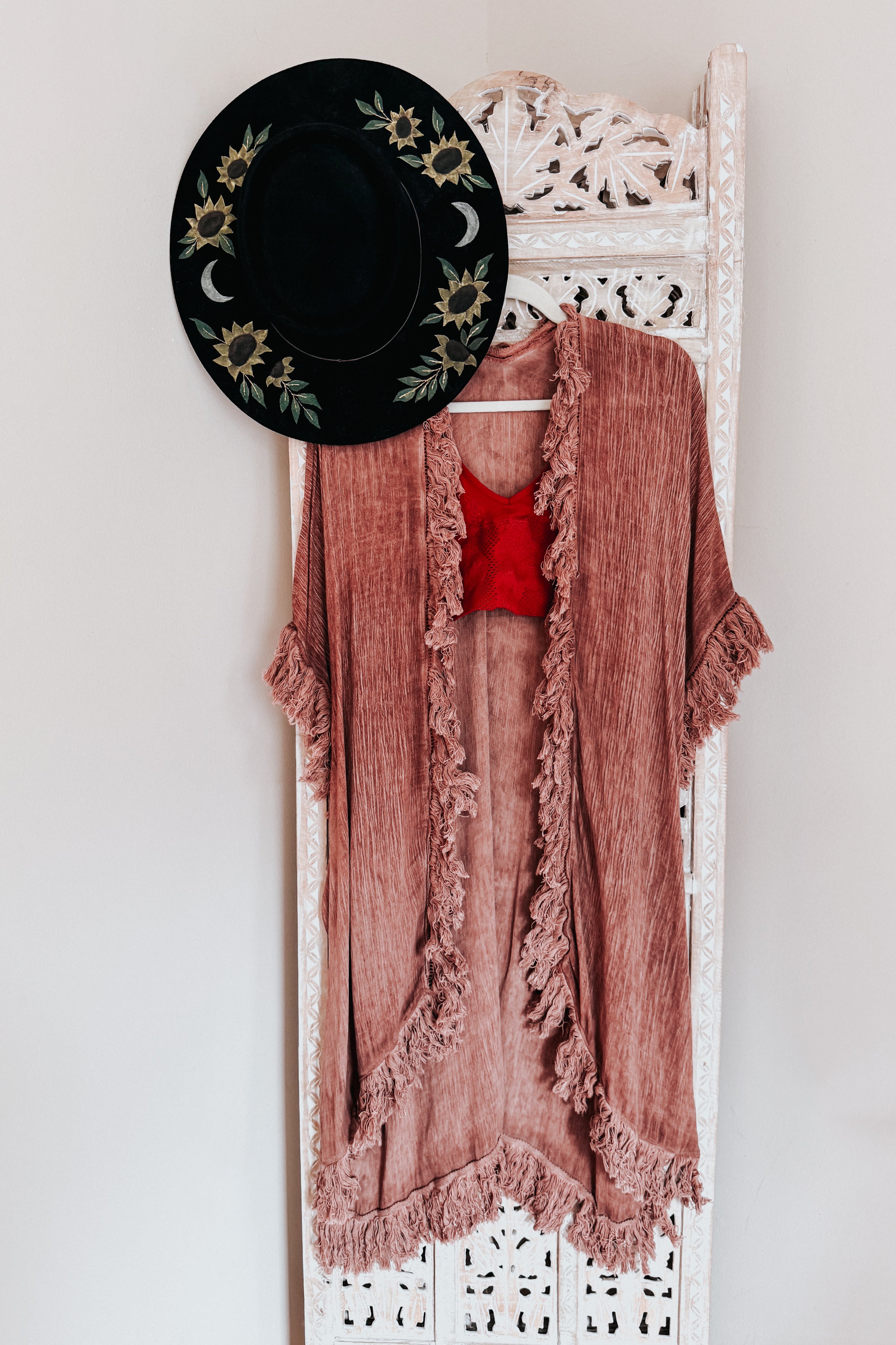 Rose-colored sunbleached duster with fringe trim hanging on ornate white wooden screen beside black floral hat