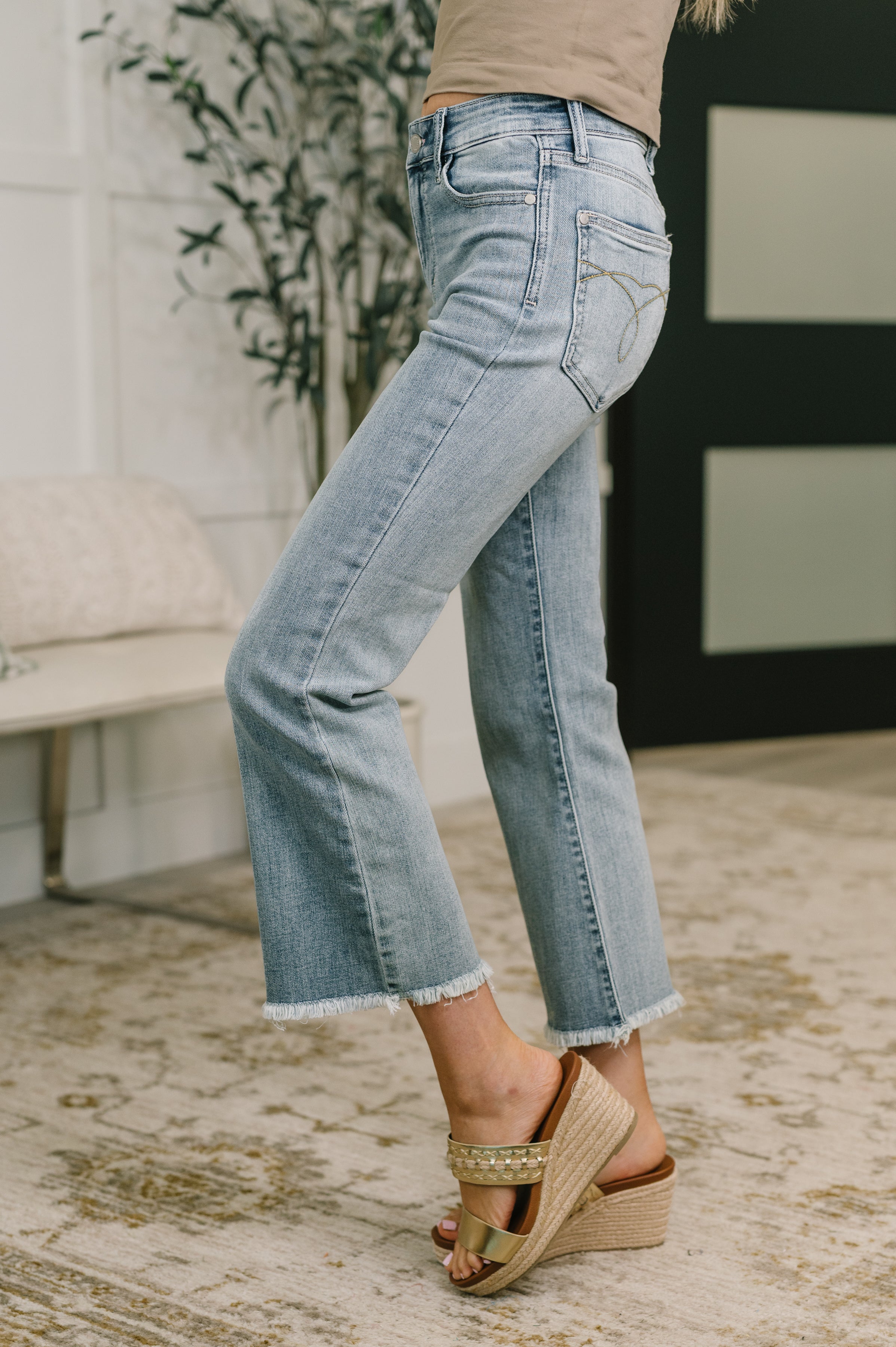 Side view of mid rise blue denim jeans with a subtle flare and frayed hem paired with gold wedge sandals indoors