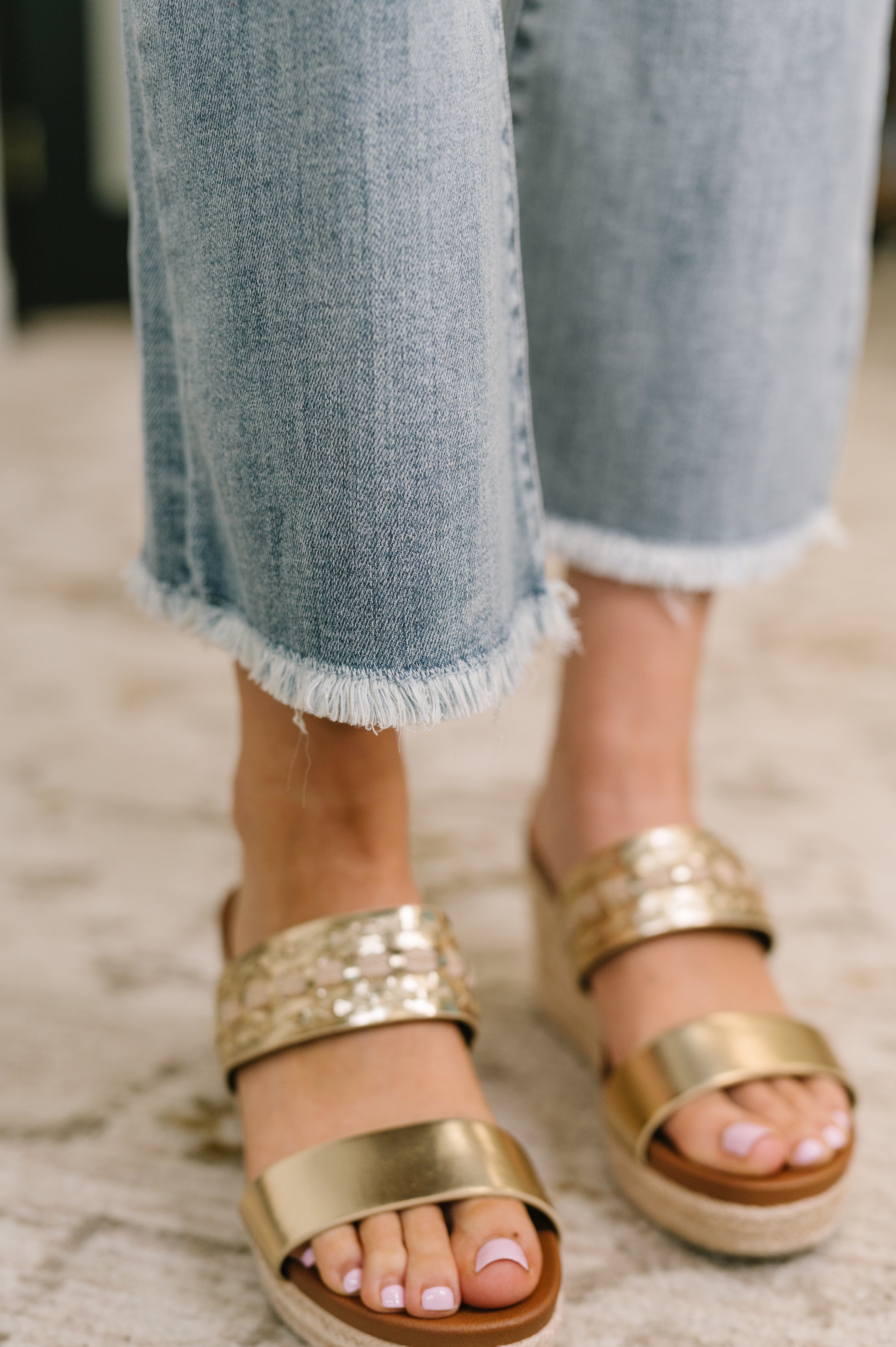 Close-up of blue mid-rise jeans with frayed hems paired with gold metallic slide sandals on carpeted floor