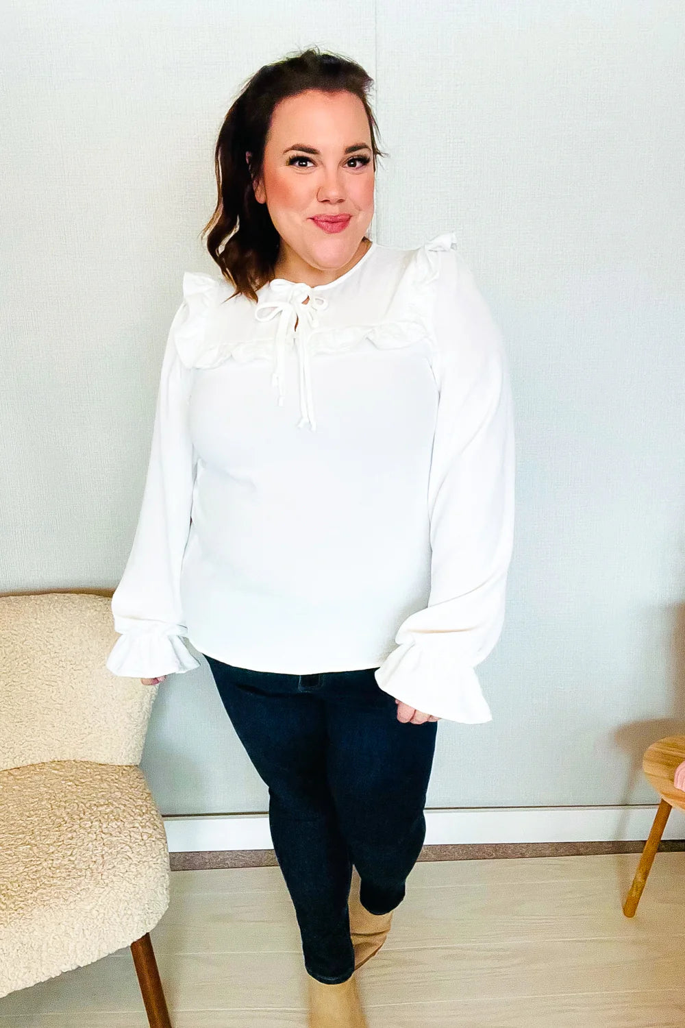 Woman wearing a white blouse with ruffled collar and tie detail, puffed sleeves with ruffled cuffs, paired with dark jeans indoors.