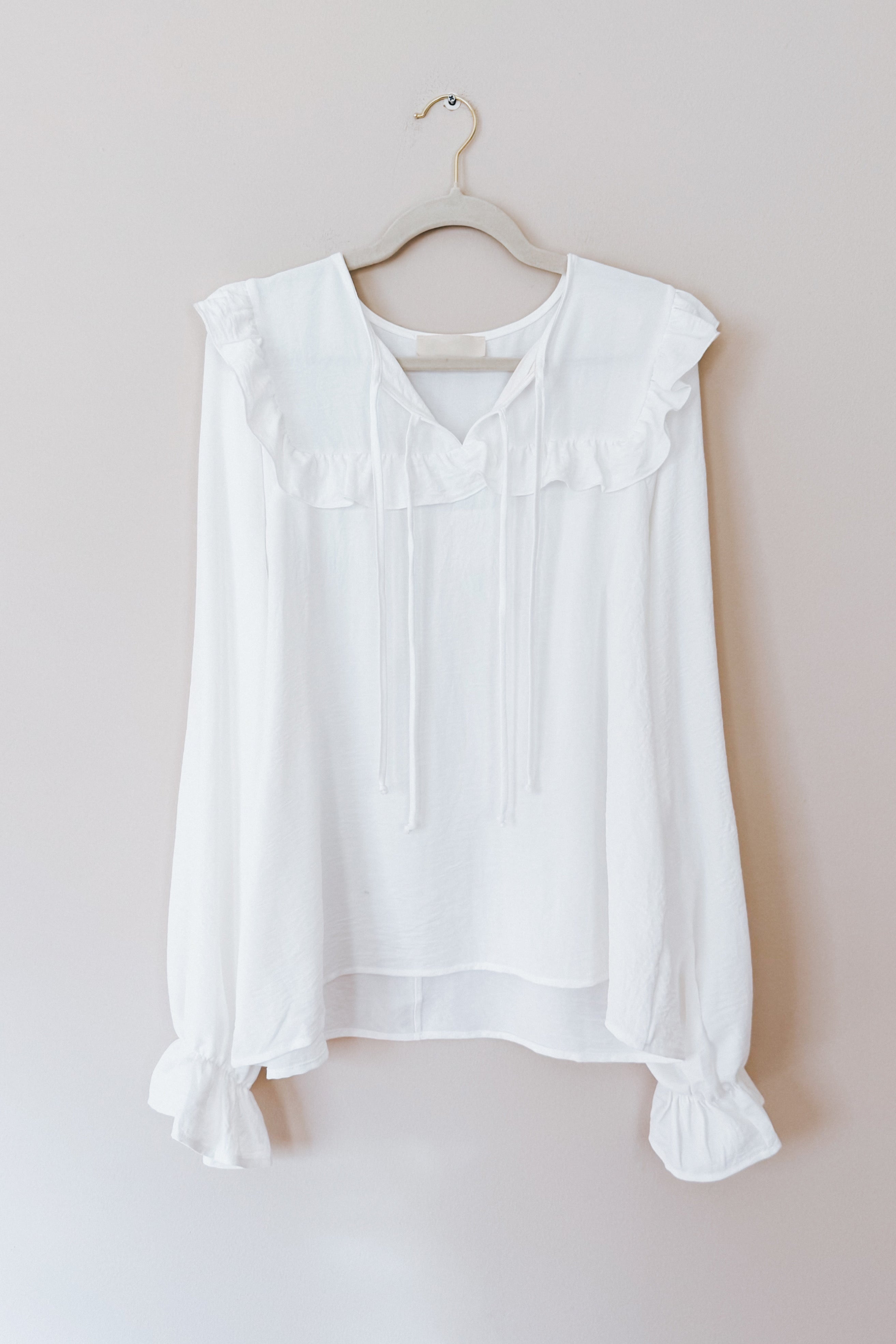 White blouse with a ruffled yoke, long bubble sleeves gathered at the cuffs, and a tie detail at the neckline hanging on a wooden hanger against a neutral background