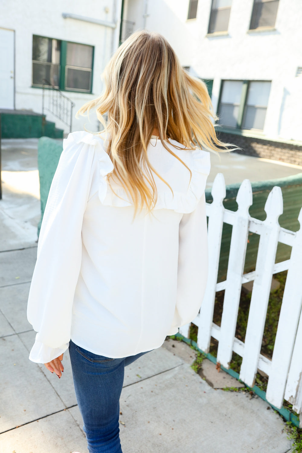 Woman with blonde hair wearing a white blouse with voluminous bubble sleeves and a ruffled back detail, paired with blue jeans outdoors near a fence.