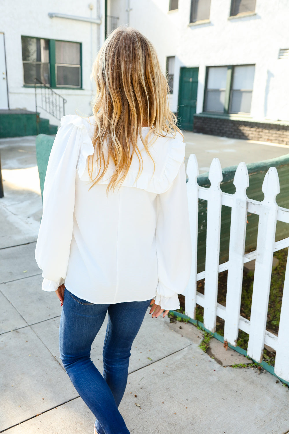 Woman wearing a white blouse with puffed sleeves and a ruffled back detail, paired with fitted blue jeans, standing near a white picket fence outdoors.