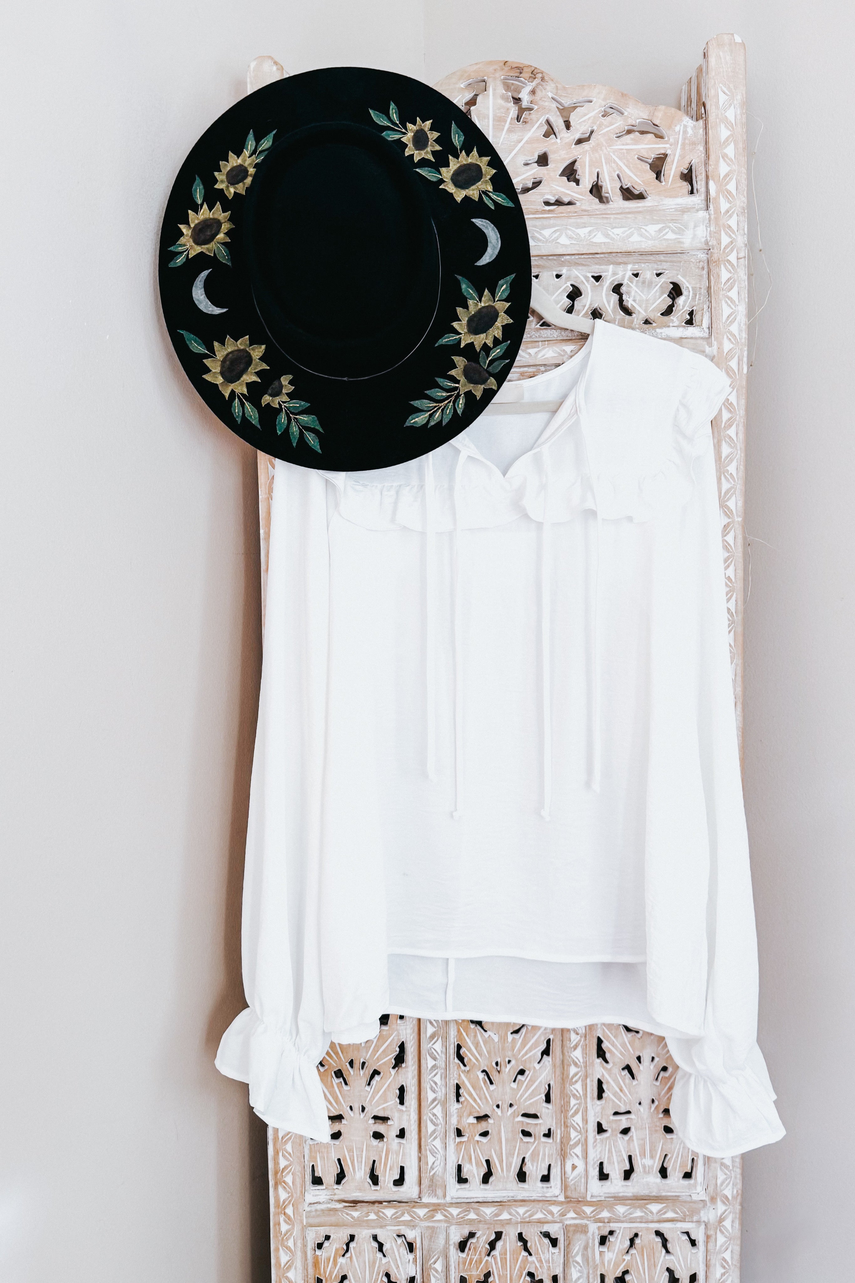 White blouse with long puffed sleeves featuring ruffled cuffs and a gathered neckline with ties, hanging on an intricately carved wooden panel next to a black wide-brim hat decorated with sunflowers and crescent moons.