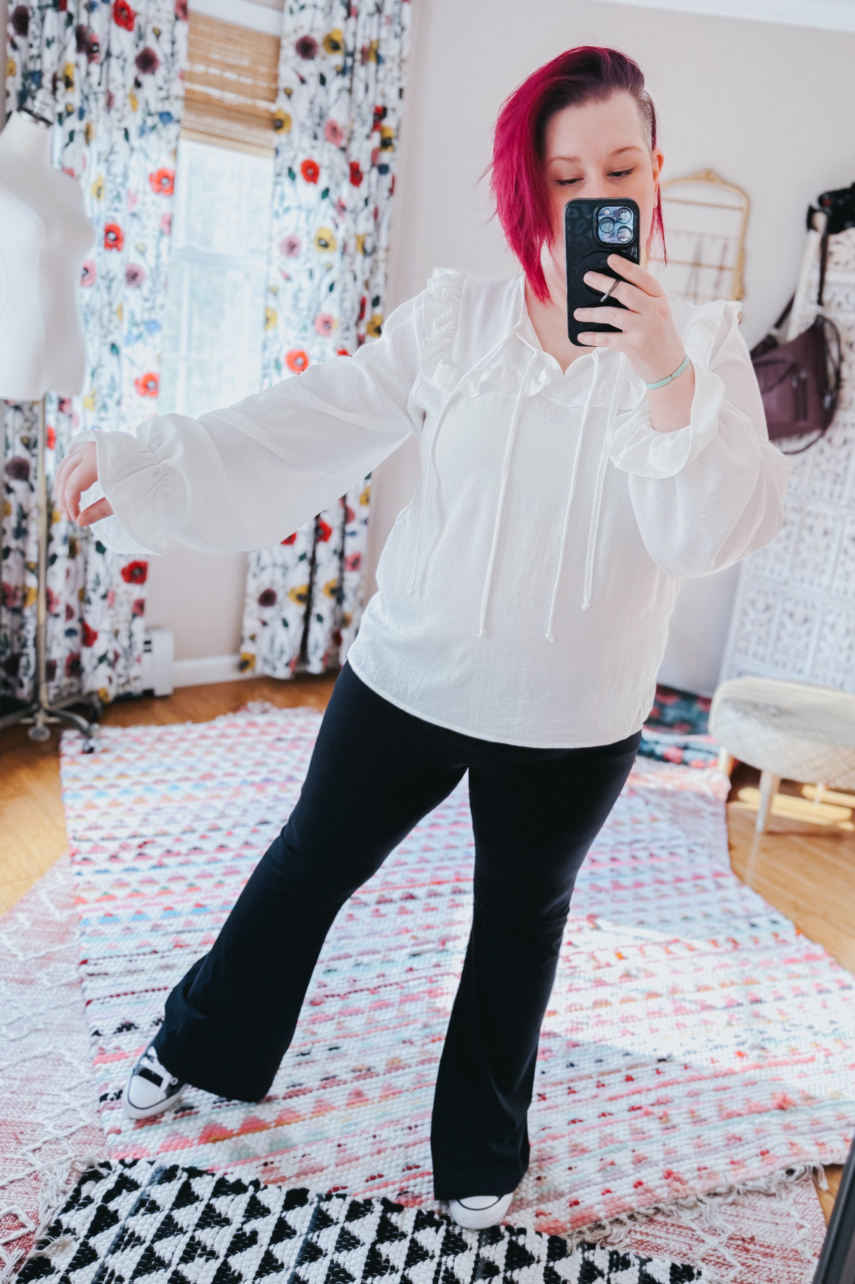 Woman taking a mirror selfie wearing a white blouse with puffed sleeves, ruffle trim, and tie details, paired with black pants indoors.