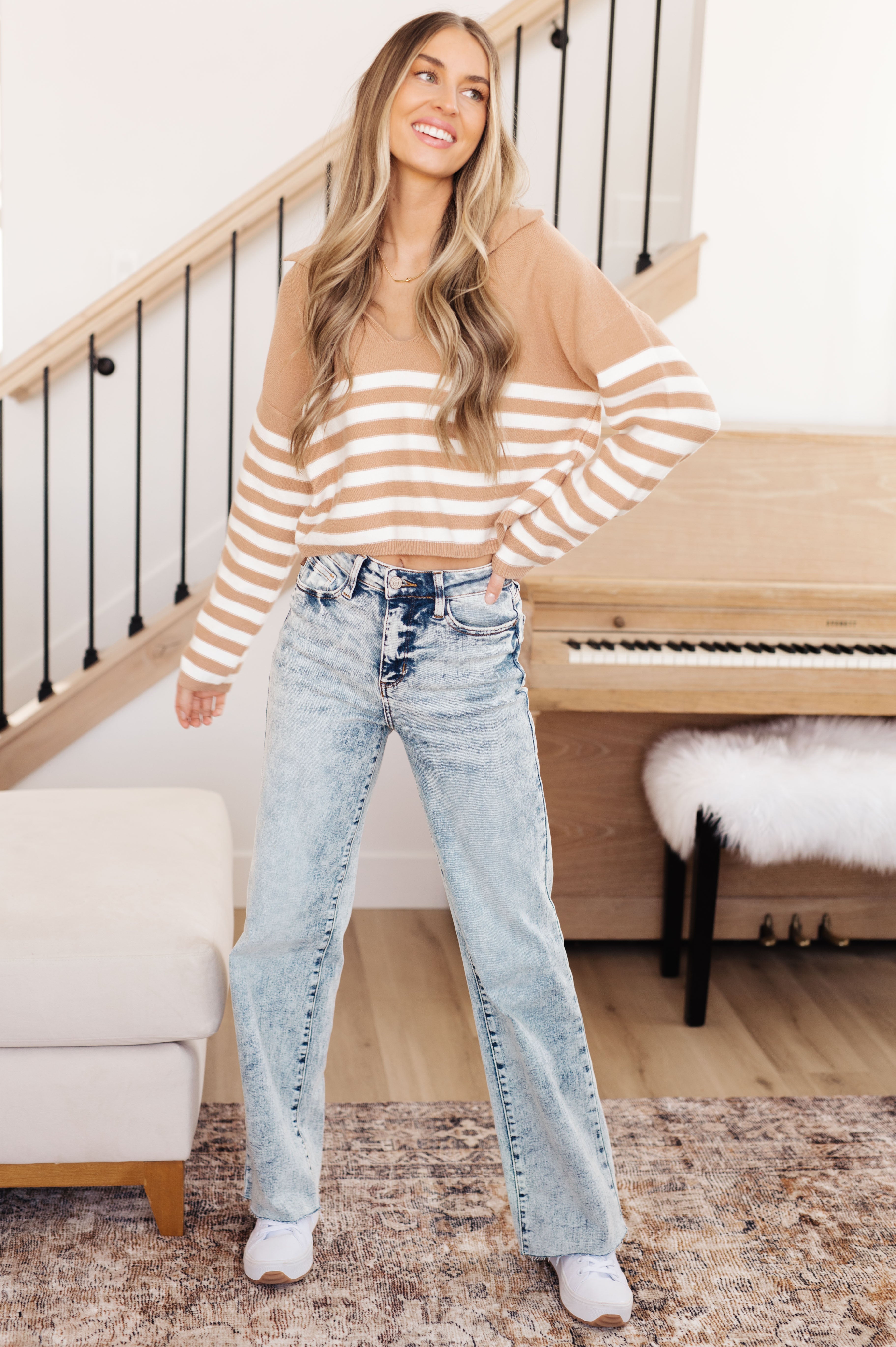 Woman wearing a mocha and white striped sweater with a relaxed fit paired with light wash high-rise jeans and white sneakers, standing indoors near a piano.