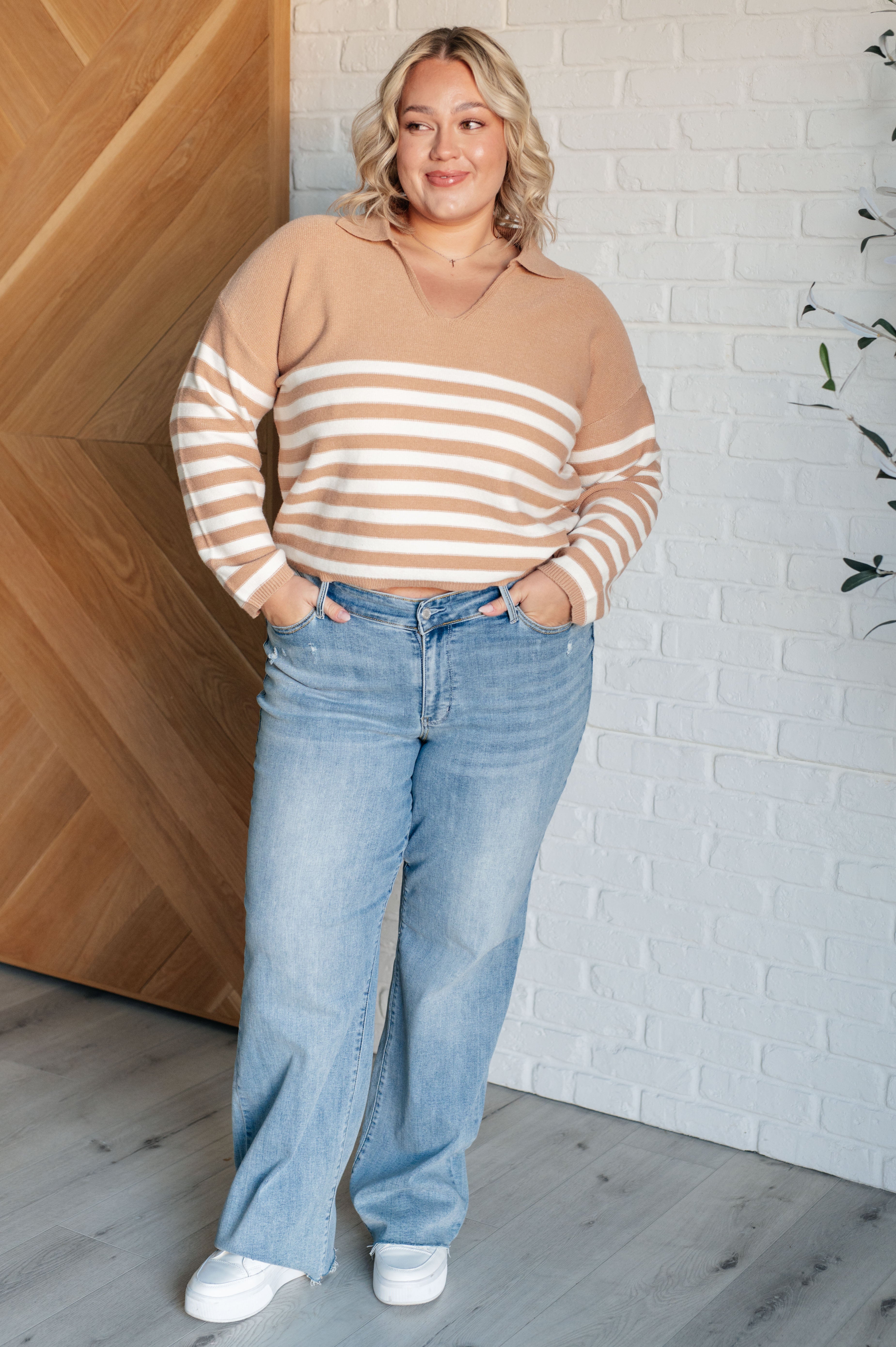 Woman wearing a mocha striped sweater with a collar and light wash high-rise jeans, standing indoors against a white brick wall and wooden door.