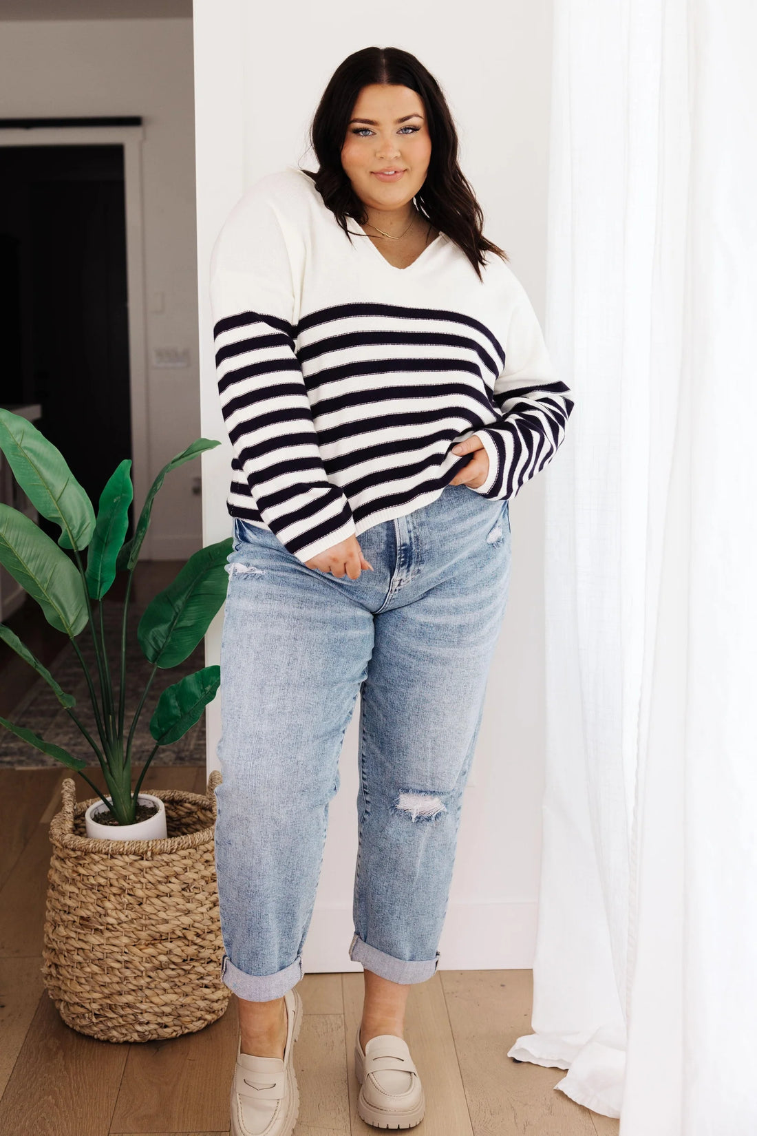 Woman with dark hair wearing a white sweater with navy horizontal stripes and light wash cuffed jeans, standing indoors near a woven basket plant.