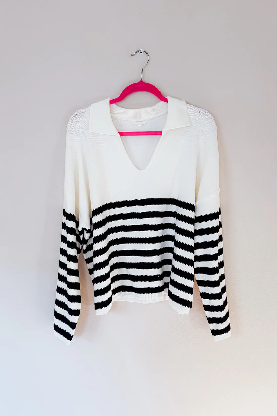 White sweater with a deep v-neck collar and bold black horizontal stripes across the lower body and sleeves, hanging on a pink hanger.