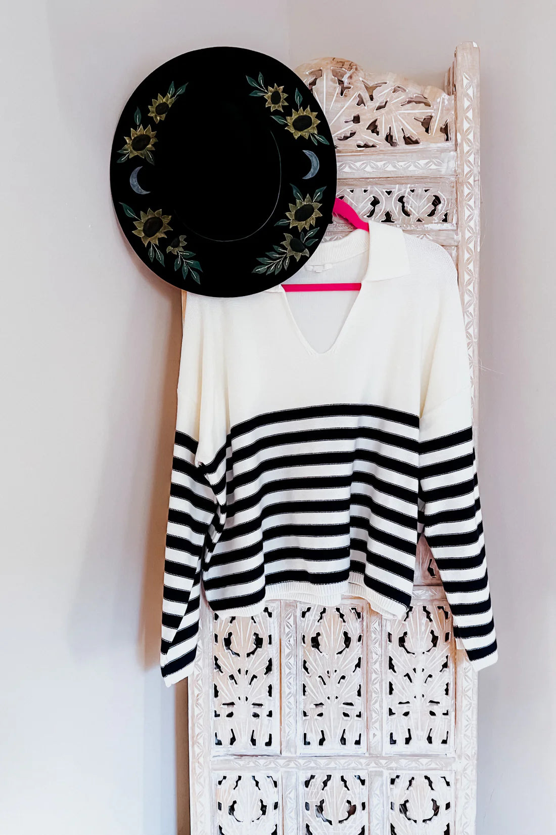 White and black horizontal striped sweater with a collar, hanging on a carved white wooden screen next to a black hat decorated with sunflowers and crescent moons.