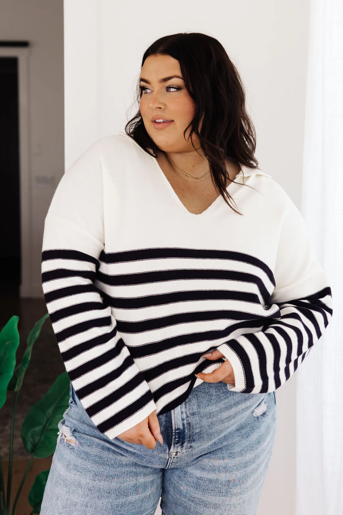 Woman with dark wavy hair wearing a white sweater with bold black horizontal stripes and distressed light wash jeans, standing indoors near a plant.