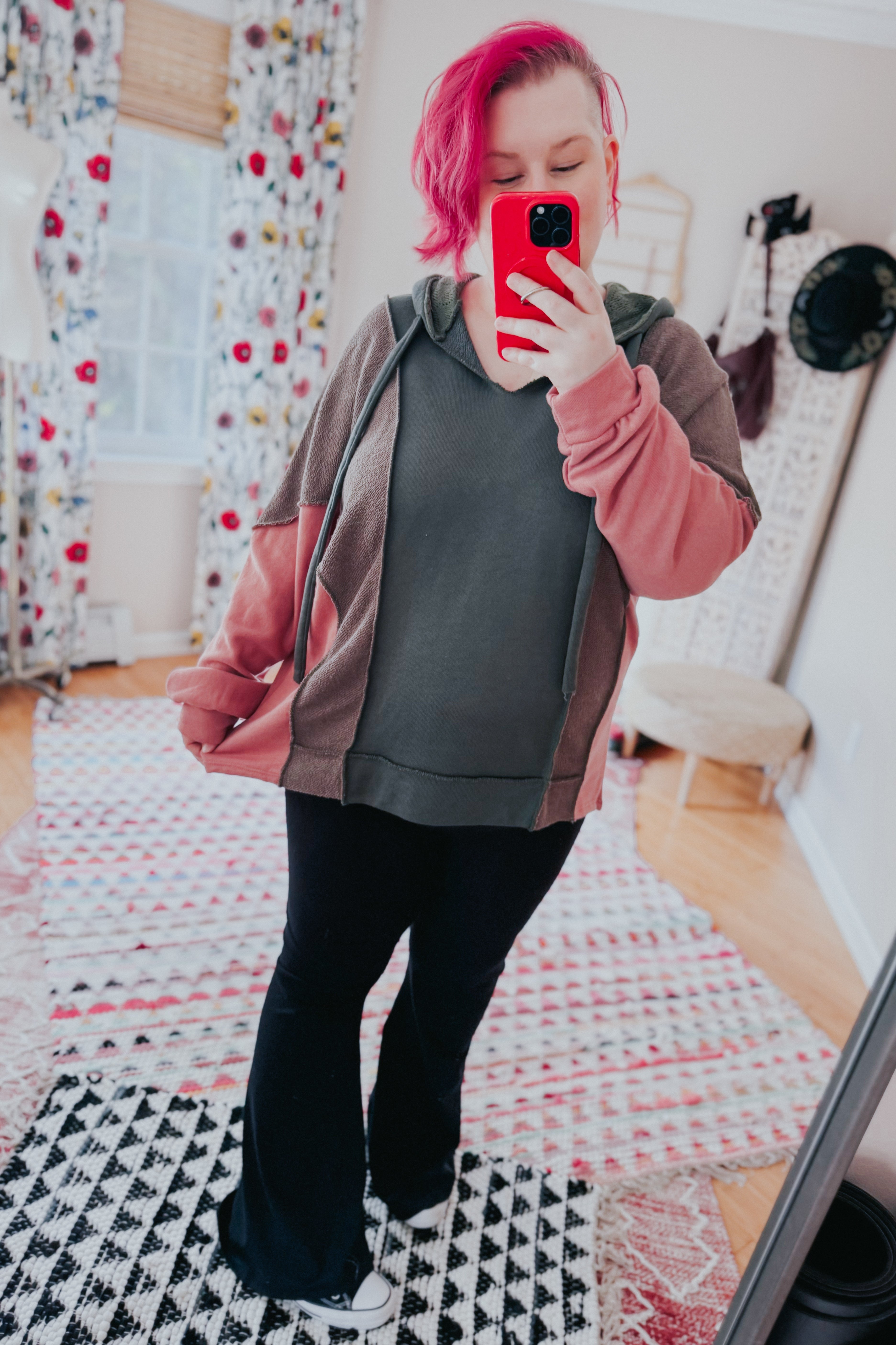 Woman with pink hair wearing a relaxed fit color block hoodie in sage, muted brown, and dusty pink taking a mirror selfie in a cozy room with patterned rugs.