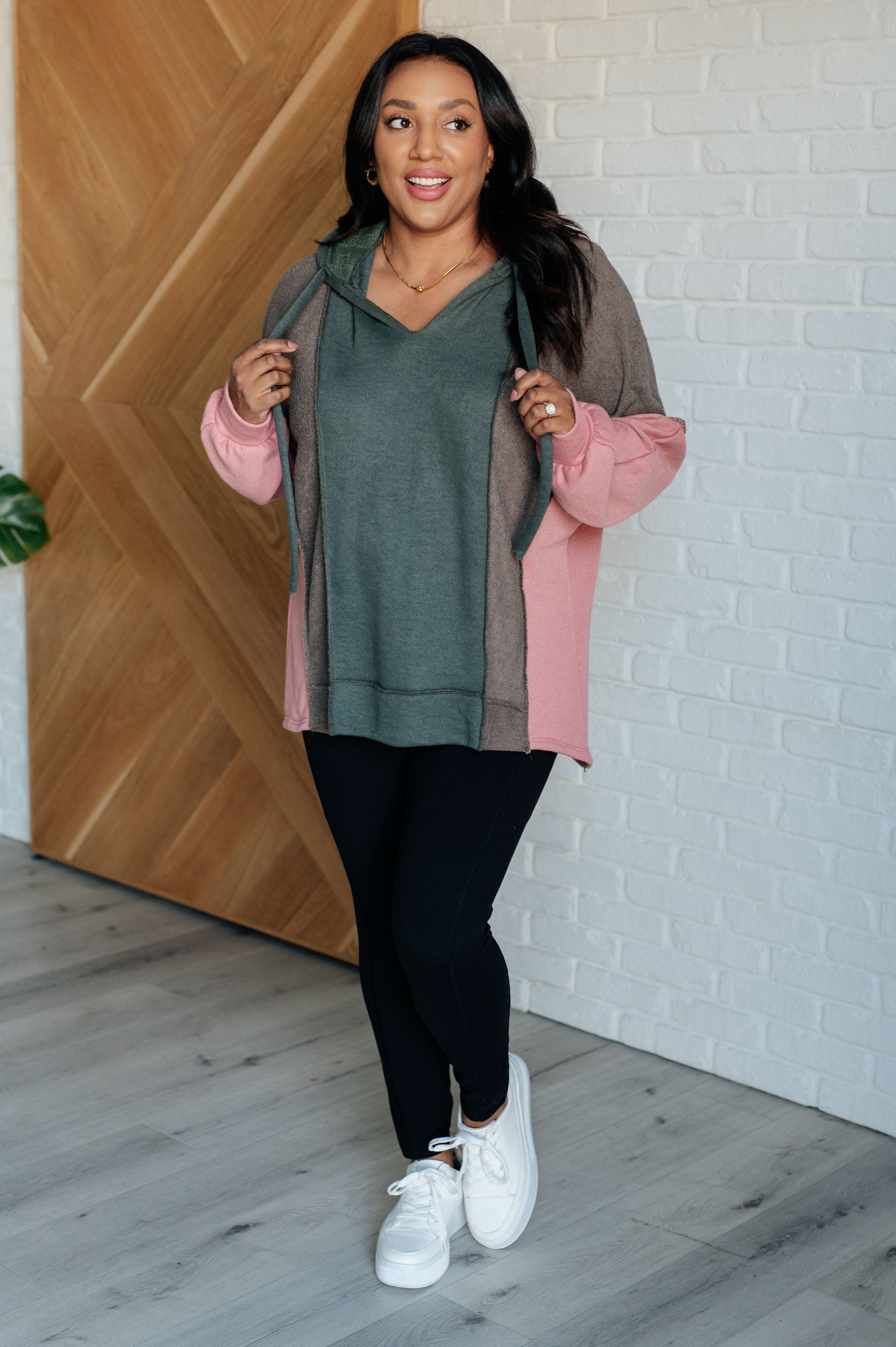 Woman wearing a relaxed fit color block hoodie in sage green, muted brown, and soft pink paired with black leggings and white sneakers, standing indoors by a wooden door and white brick wall.